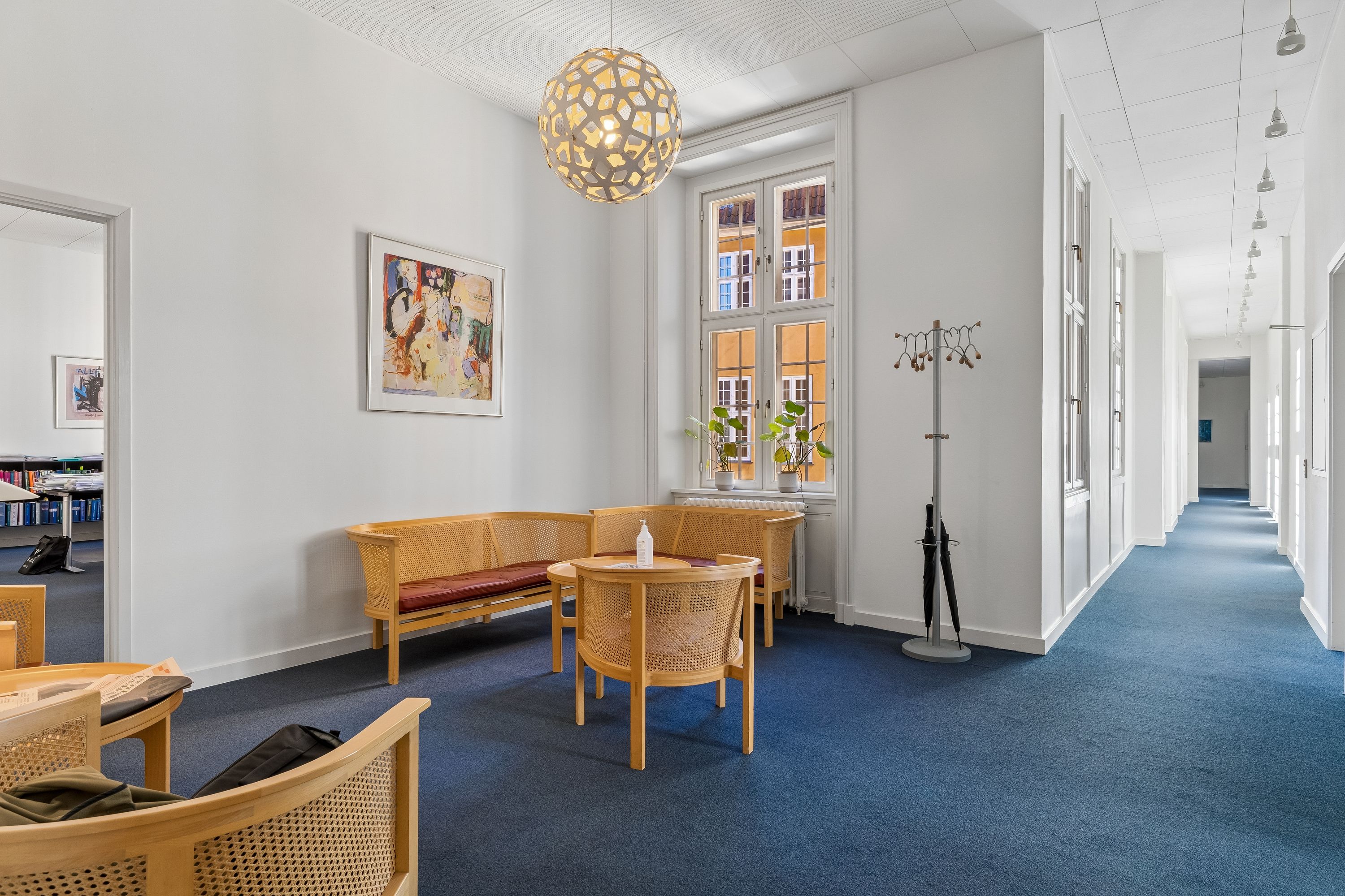 Kontor med blå gulv, lyse vægge og lysekroner. Et skrivebord med to stole og en sofa med bord i midten. Et tøjstativ med paraply og en plante i vinduet.