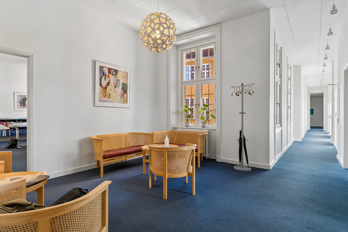 Kontor med blå gulv, lyse vægge og lysekroner. Et skrivebord med to stole og en sofa med bord i midten. Et tøjstativ med paraply og en plante i vinduet.