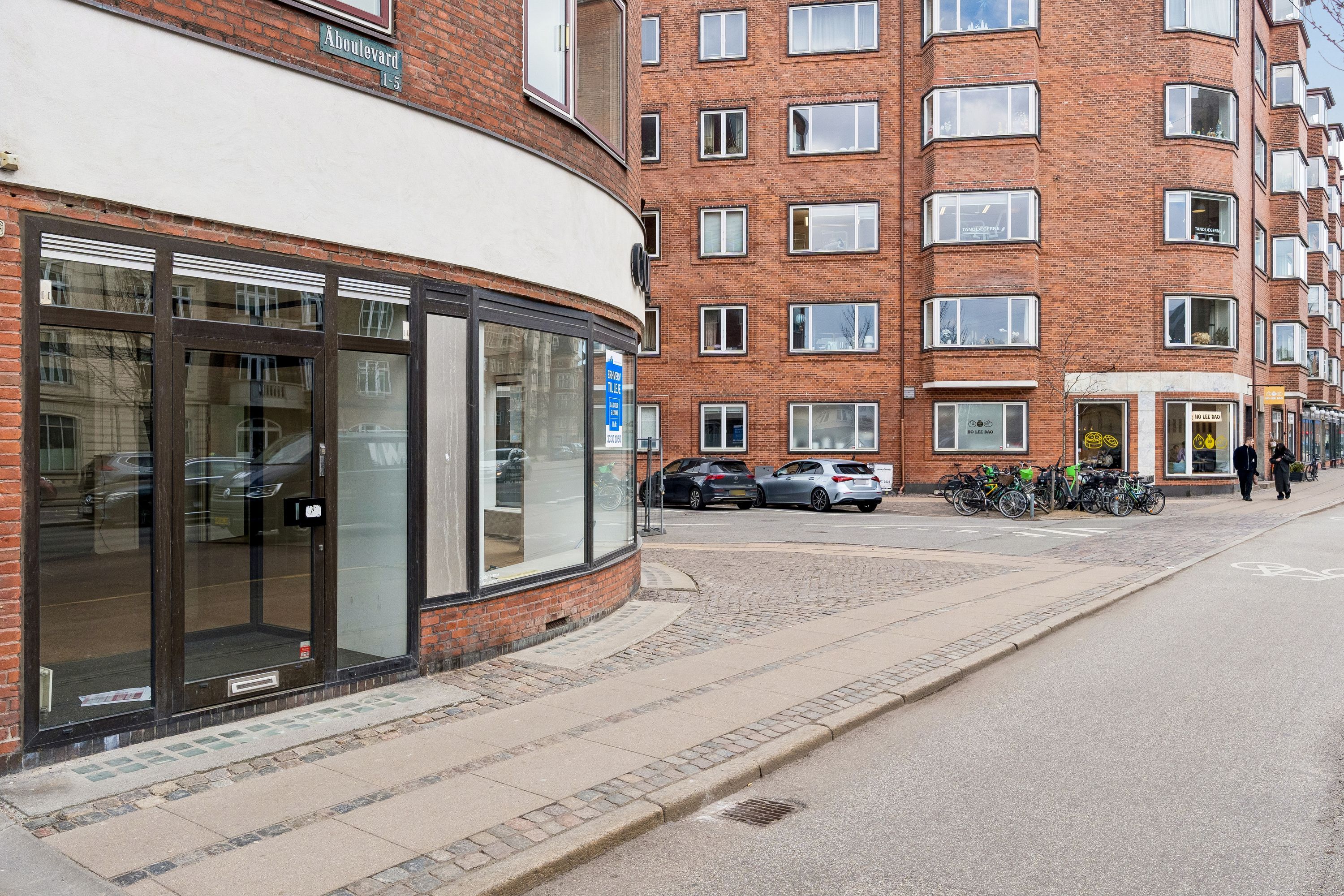 Hjørnebygning med glasvinduer og røde murstensvægge langs Åboulevard. Parkerede biler og cykler langs fortovet. To mennesker i forgrunden og adskillige vinduer på baggrunden.