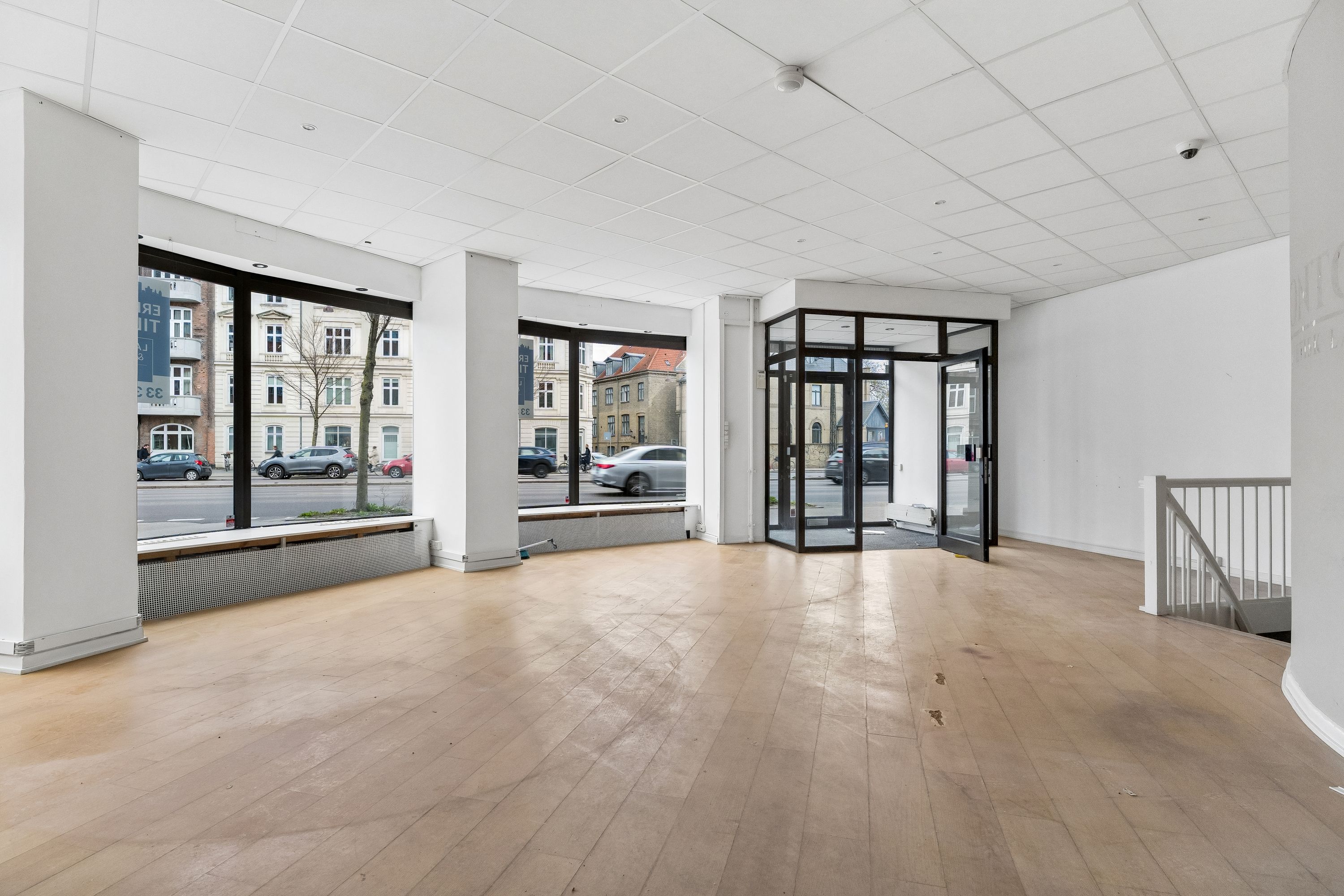 Moderne butik med store ruder og åbne døre mod gade med biler og et rødt bilnummerplade. Stående radiator, høj hvid gulv og høj hvid loftsbeklædning.