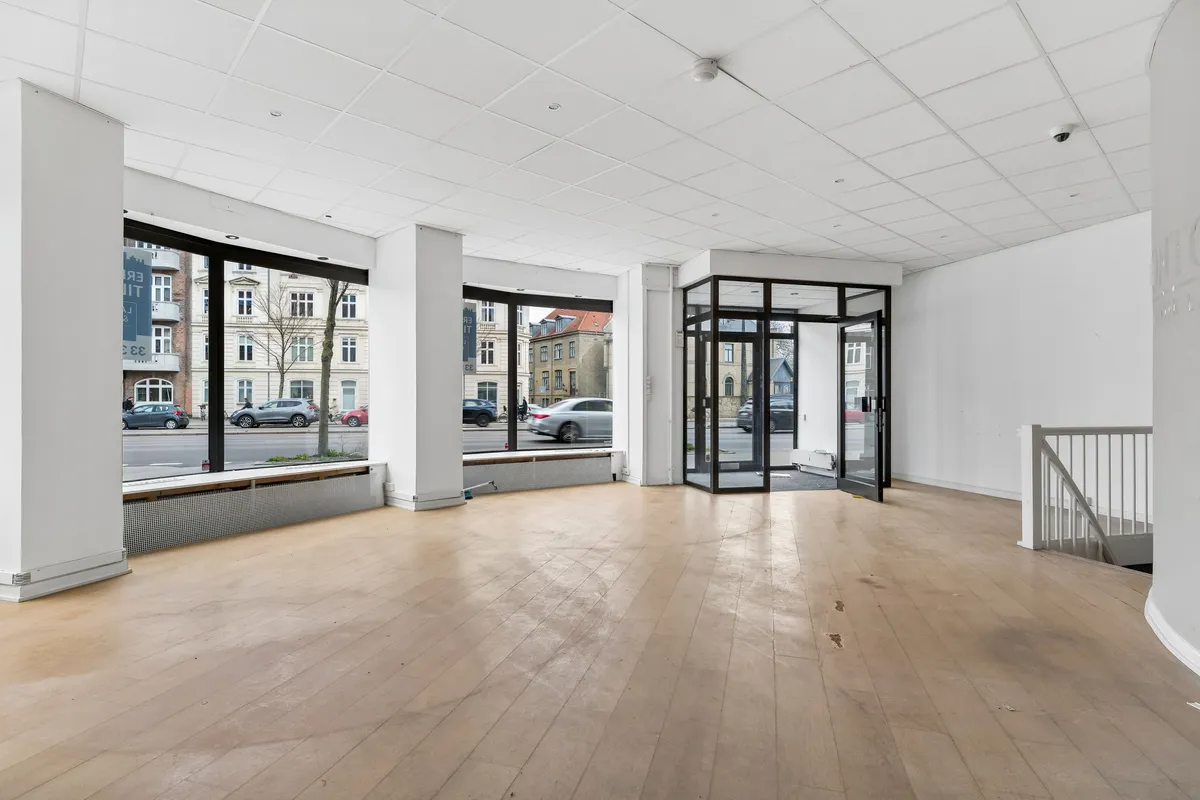 Moderne butik med store ruder og åbne døre mod gade med biler og et rødt bilnummerplade. Stående radiator, høj hvid gulv og høj hvid loftsbeklædning.