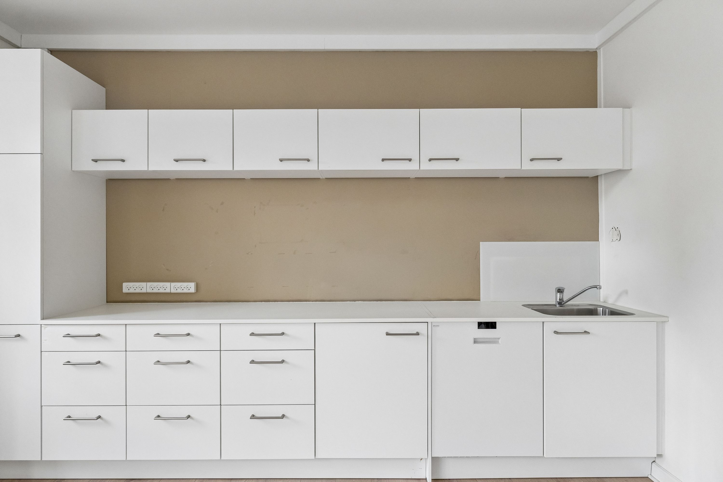 En ren og funktionel køkkeninteriør med hvide skabsmøbler og hvide vægge, en enkelt høj køleskab og en stor rustfri stål vandhane placeret over en dyb, fladt skåret rustfrit stål vaskebækken