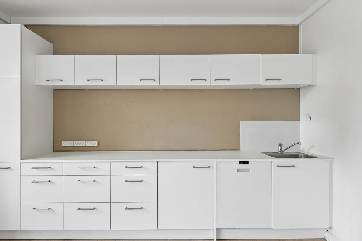 En ren og funktionel køkkeninteriør med hvide skabsmøbler og hvide vægge, en enkelt høj køleskab og en stor rustfri stål vandhane placeret over en dyb, fladt skåret rustfrit stål vaskebækken