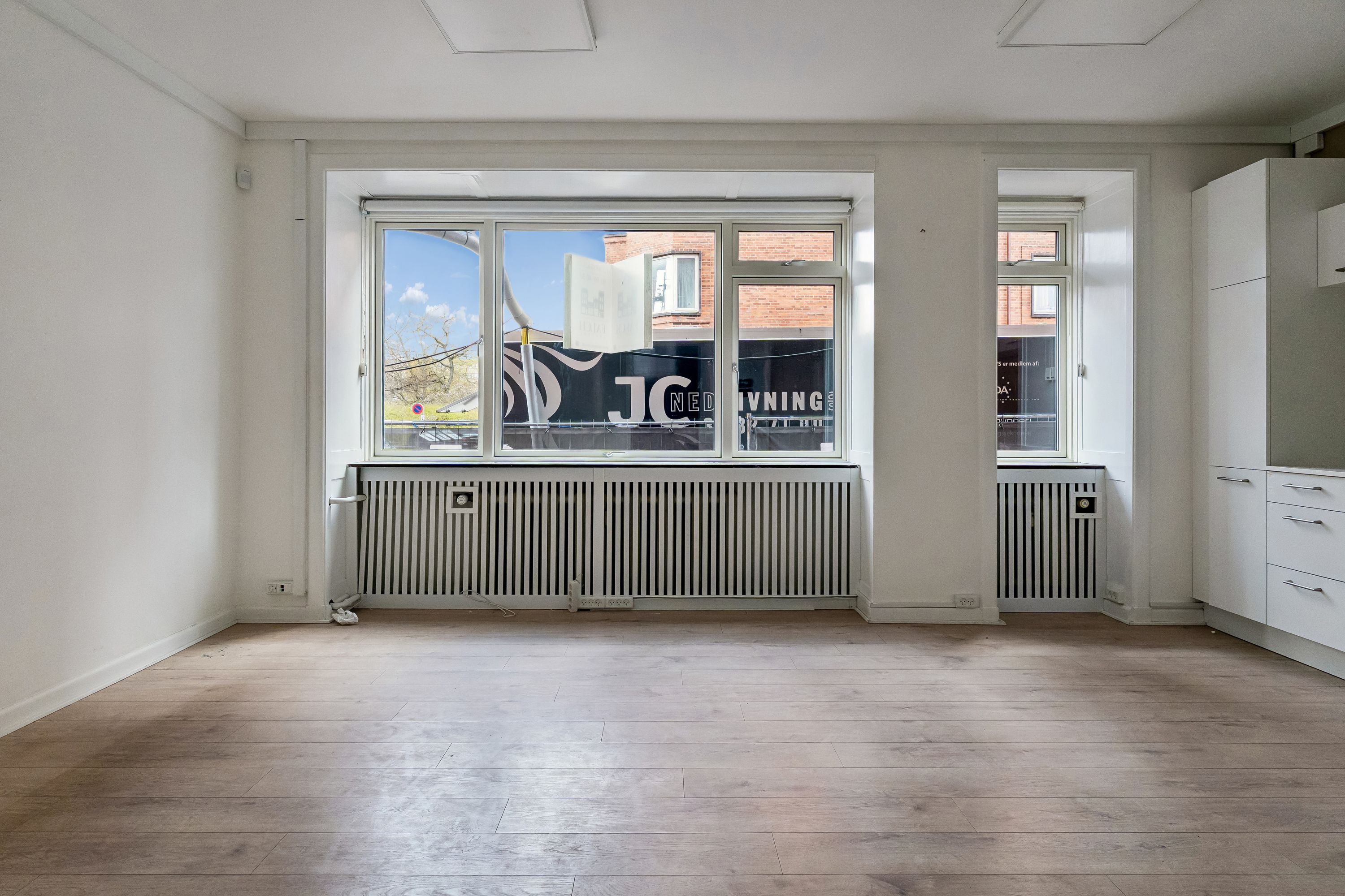 En stor stue med hvid væg og gulv, radiatorer under vindue og en stor vinduesfront ud til gaden