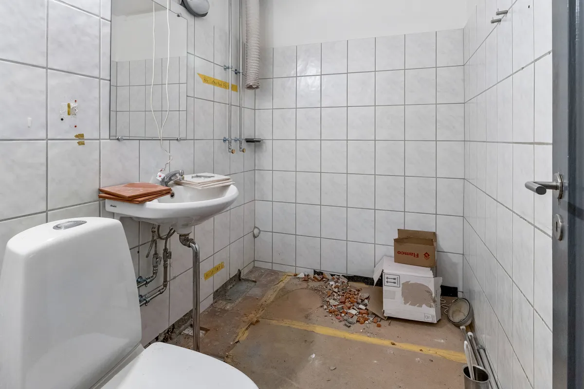En lille toiletrum med hvide fliser, en hvid toilet, en hvid vask og en metalbøjle med en sæbeholder. Et lagerkarton står ved siden af, og der er flere smadrede sten på gulvet.
