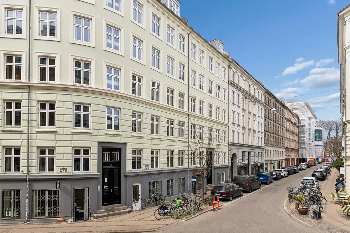 Karakteristisk lejemål med vinduer og parkerede biler langs gaden i København. Mærkeligt bygning med to indgange, flere cykler og stolpe samt høj bygning med solpaneler