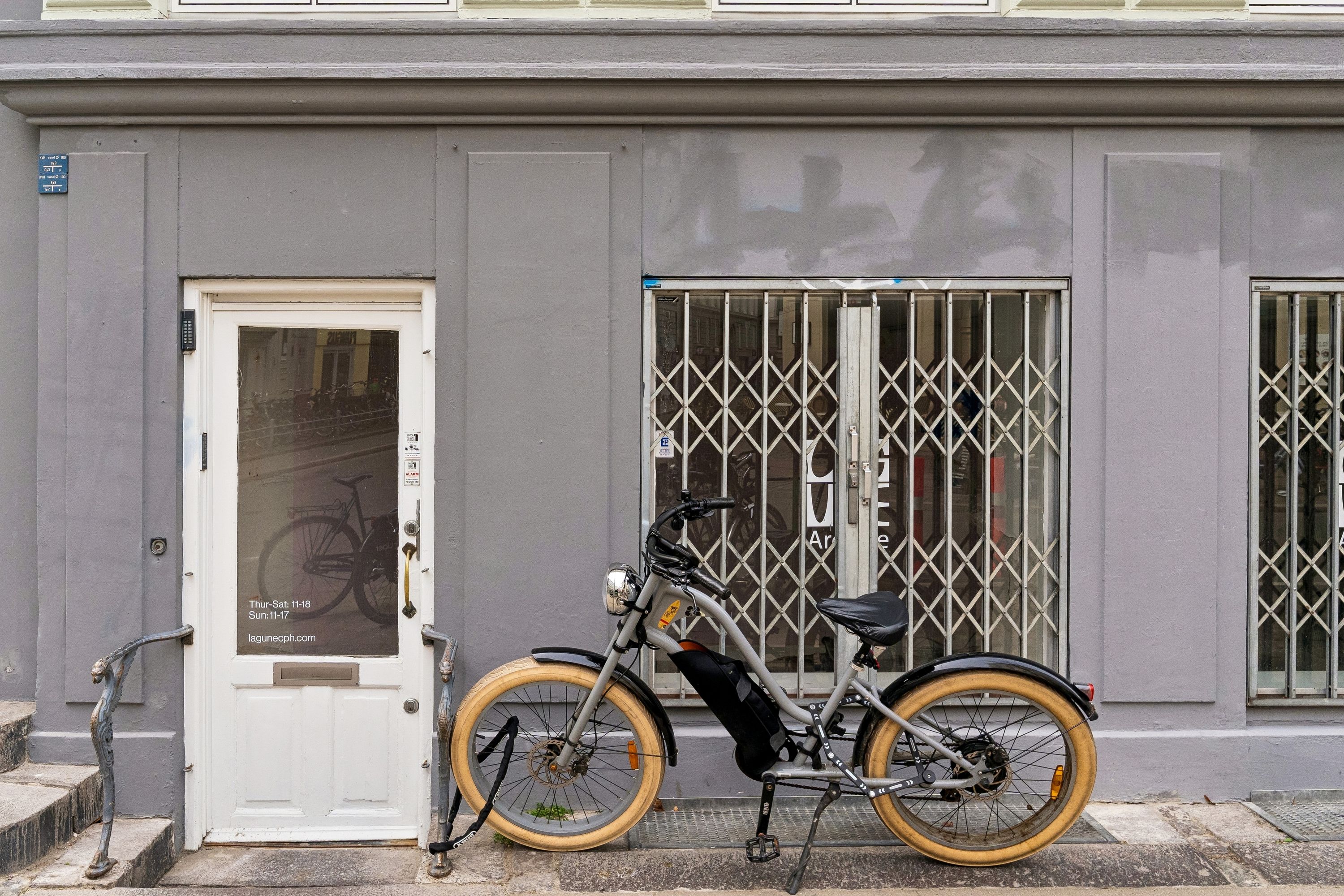 En elektrisk cykel parkeret foran en lejelejlighed med åbne jaluser og et hvidt dørpanel med tekst på glasdøren