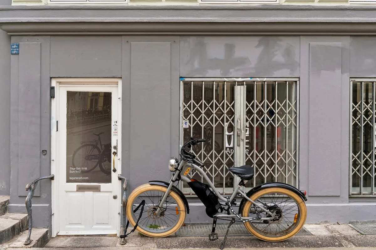 En elektrisk cykel parkeret foran en lejelejlighed med åbne jaluser og et hvidt dørpanel med tekst på glasdøren