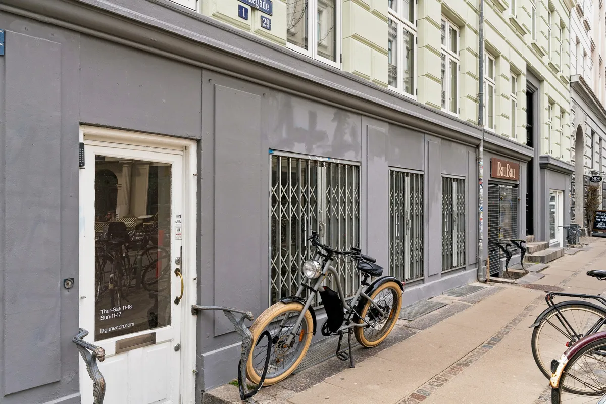 Kæde udenfor en butik på Birkegade 1, København. Butikken er låst med en metaldør og der er tre cykler parkeret udenfor. Et schild med tekst er hængt op i vinduet