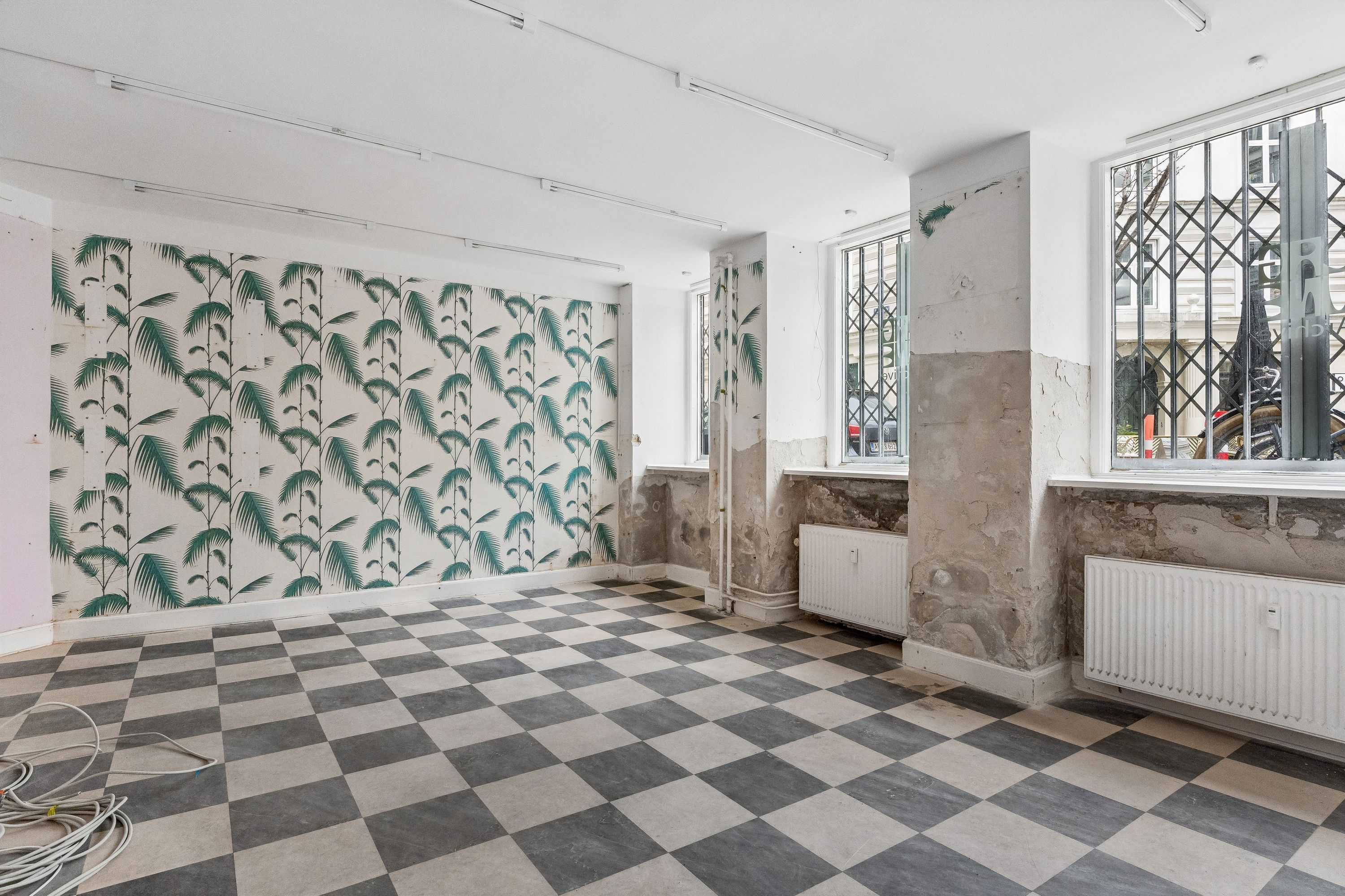 Køkken med krydsfinergulv og væg med palmetræ tapet. Køkken med radiator og en dør til siden. Væg med hængende lysekroner