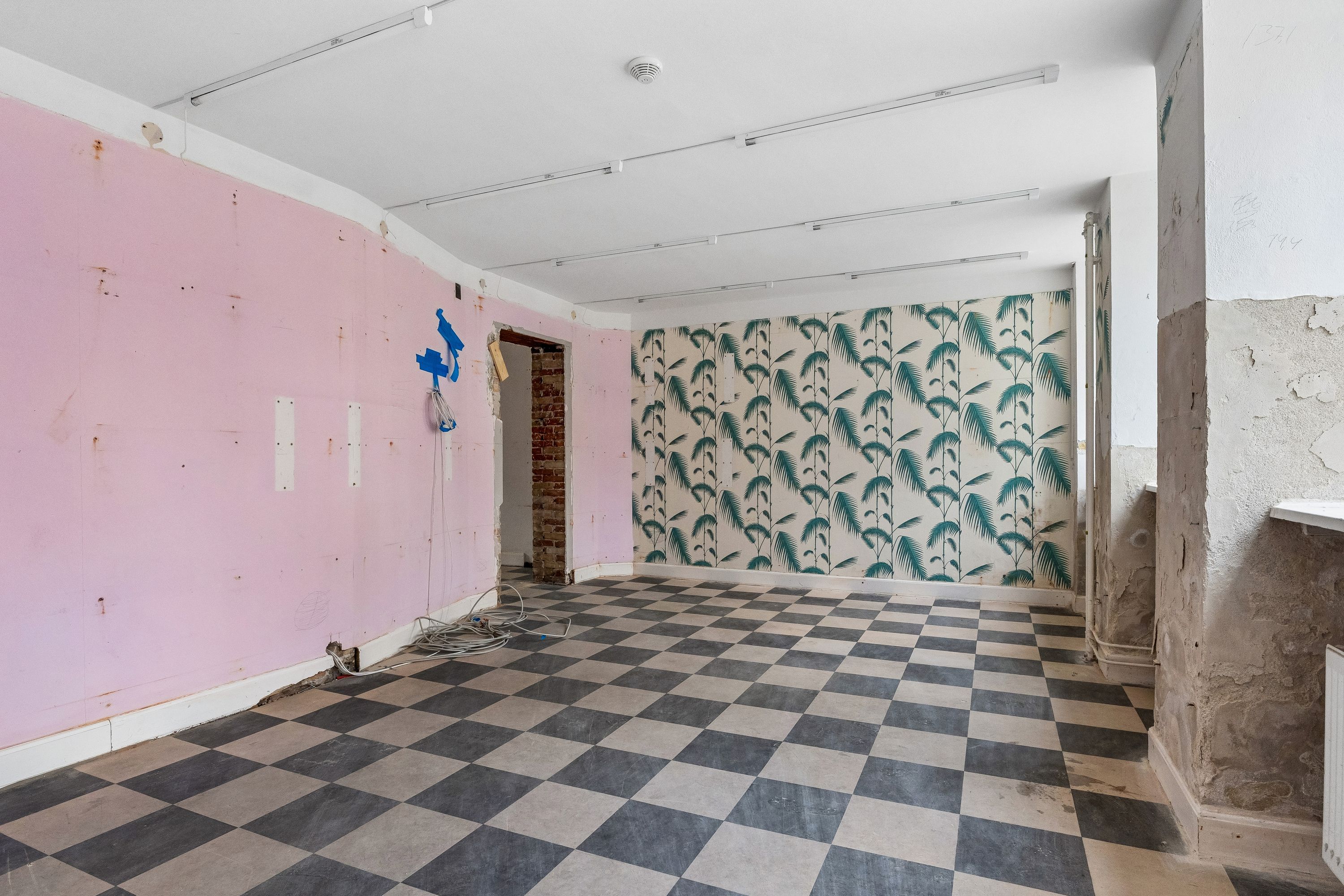 Tomt rum med glat gulv og lyse og mørke firkantede fliser, en lysgul væg og en væg med mønstret tapet, lyse og mørke loftslampe og et sølvfarvet radiator