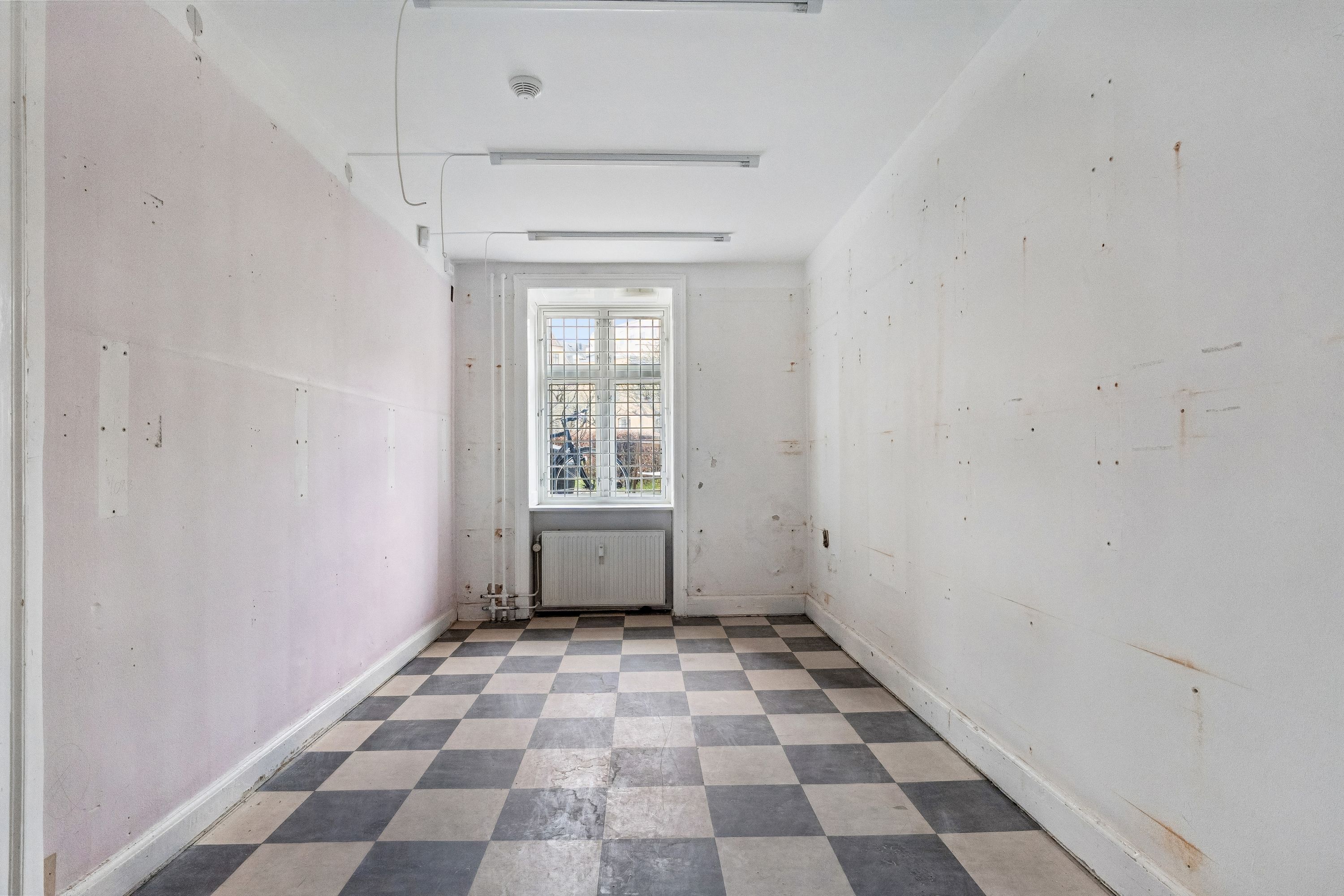 Kort og længe korridor med stribet gulv og hvide vægge med flere pletter og huller i væggene og en hvid radiator under vinduet