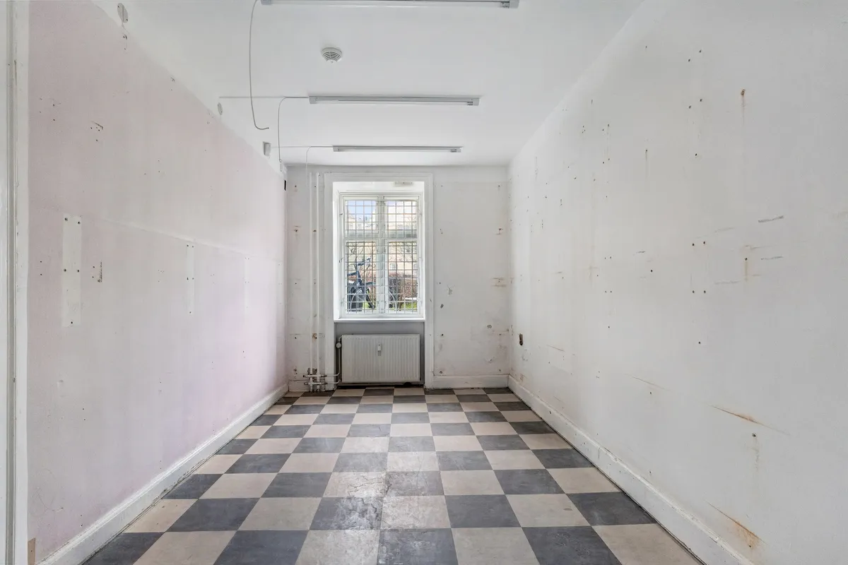 Kort og længe korridor med stribet gulv og hvide vægge med flere pletter og huller i væggene og en hvid radiator under vinduet