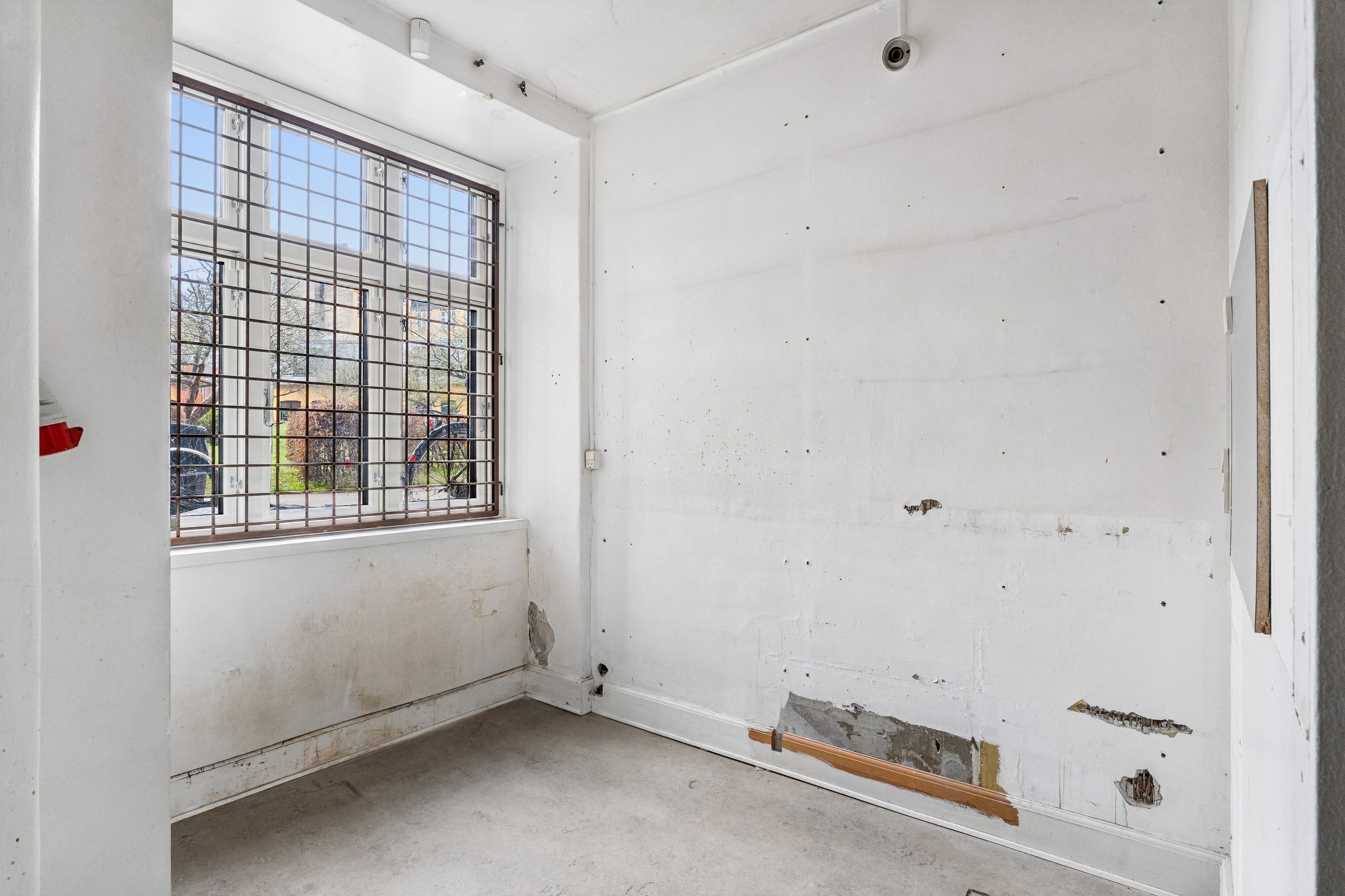 Rum med tomme hvide vægge og glasvindue med metalrist. Der er flere huller og revner i væggene. Der er et rødt objekt hængende på en af væggene.