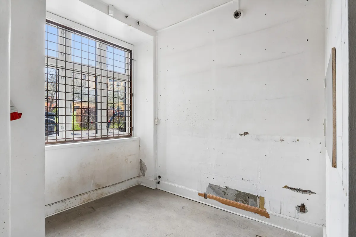 Rum med tomme hvide vægge og glasvindue med metalrist. Der er flere huller og revner i væggene. Der er et rødt objekt hængende på en af væggene.