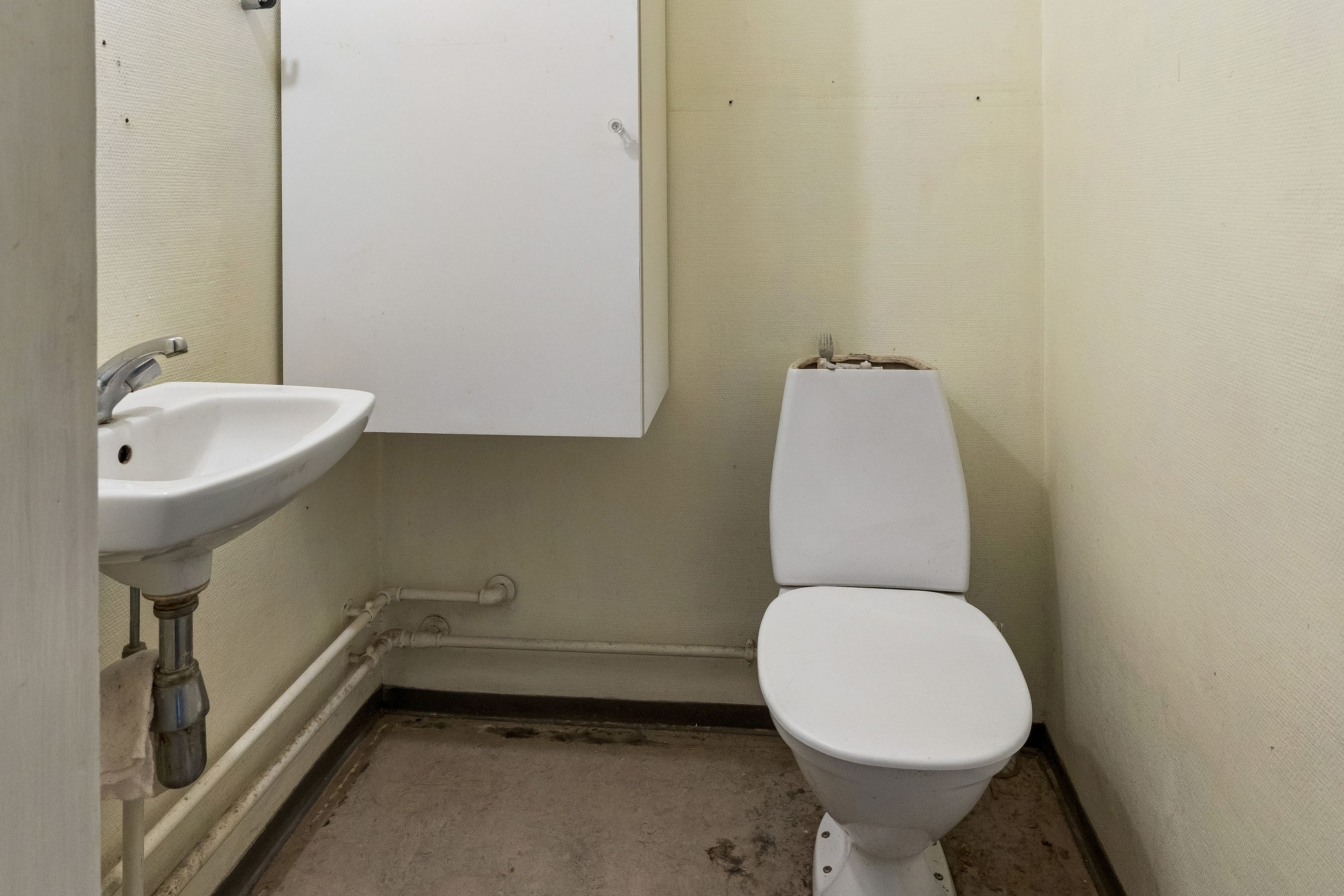 En lille toiletrum med en hvid mur, et toilet med lukkelåg og en hvid vask med vandhane