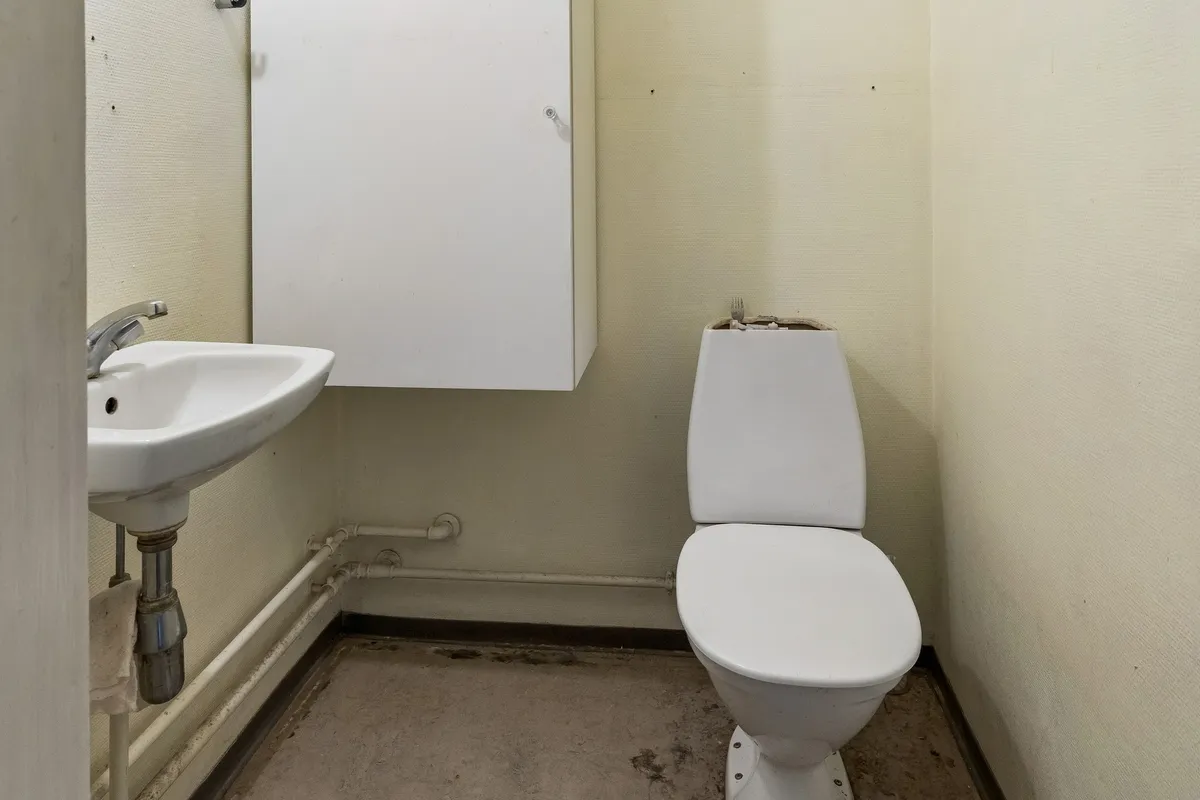 En lille toiletrum med en hvid mur, et toilet med lukkelåg og en hvid vask med vandhane