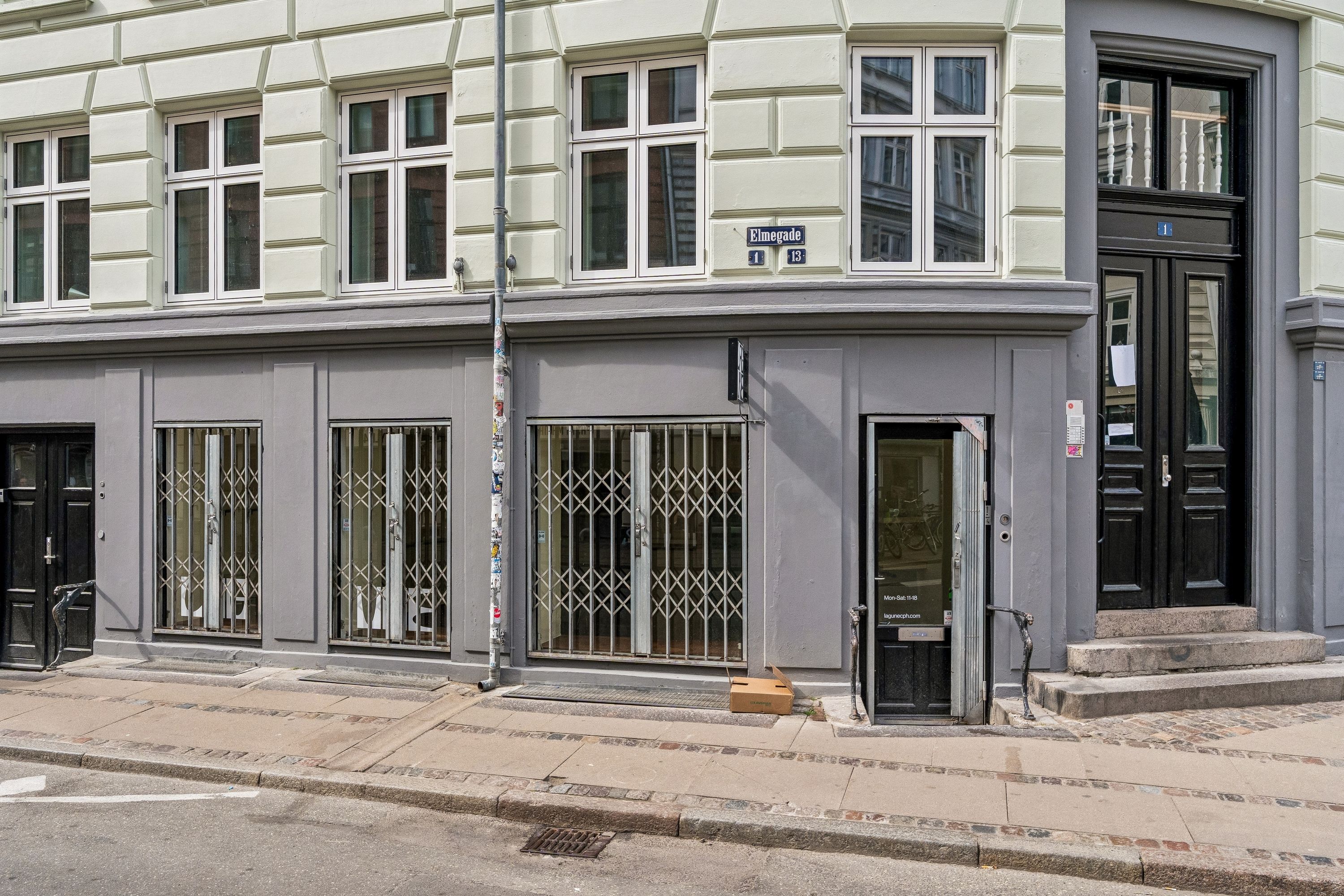 Butikstilbud på Birkegade 1 med store glasruder og lukket rollegade. Styrke metalruder og skrifttavle med adressen Elmegade 13. Sort trætrappe med skraldkasse til højre