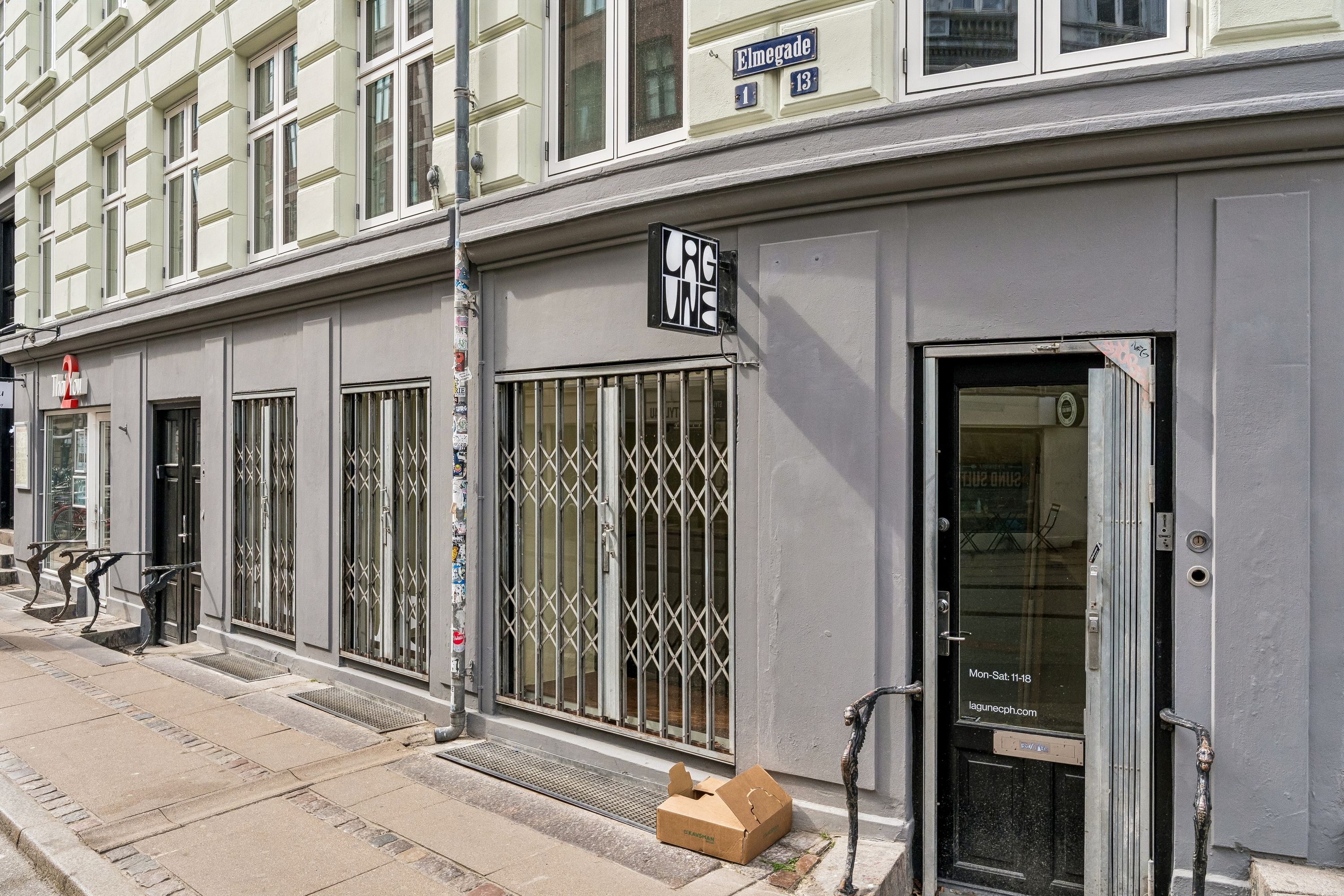 Regnbuefarvet facade med glasvinduer og lagerdør. Stående kasse med åben dør og skriftlig information om åbningstider. Ståbord, sammen med skraldespand og gul tekst på facaden