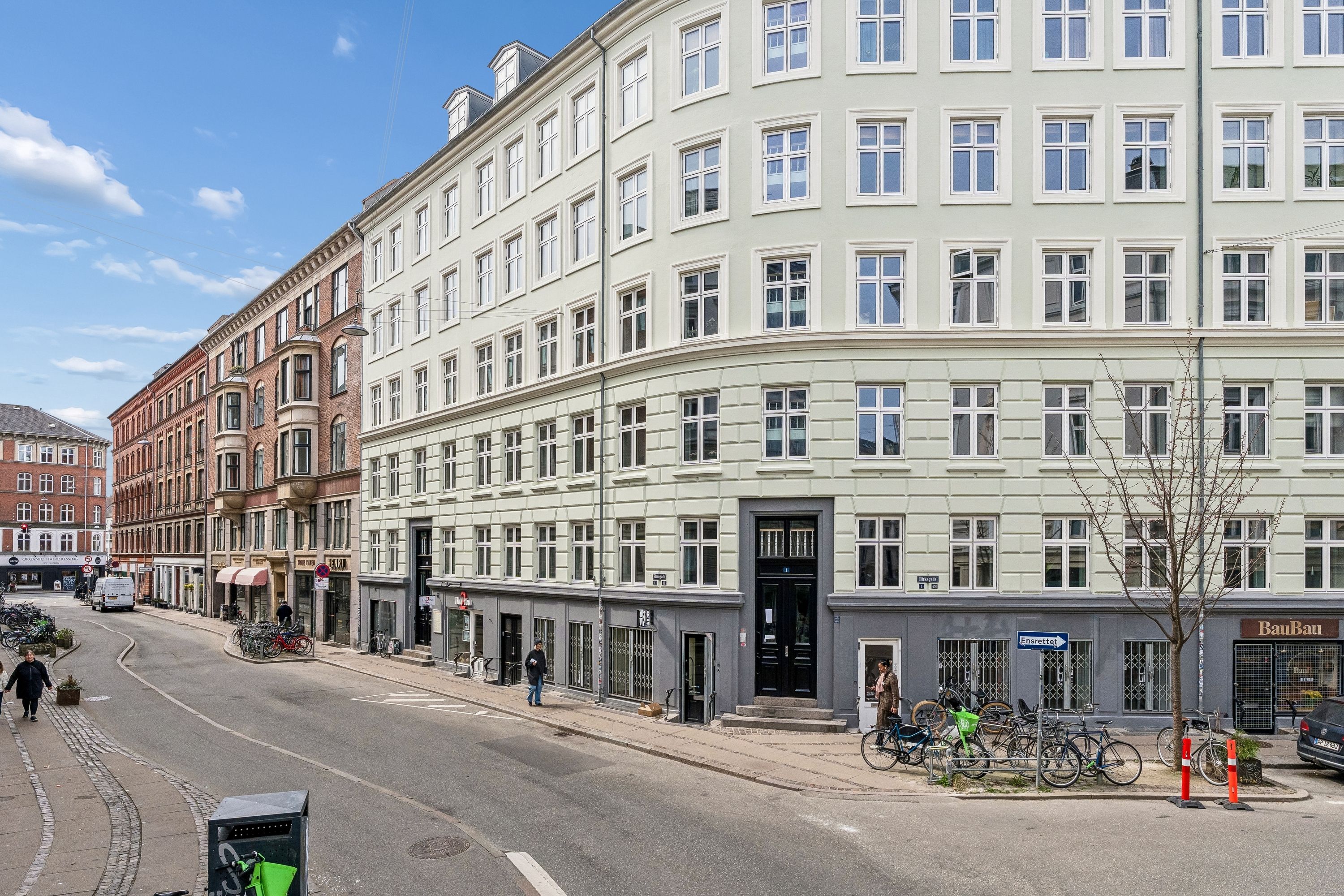 Forretningslokal med mange vinduer på Birkegade i København. Folk går rundt, og der er cykler parkeret foran. Solskin og blå himmel.