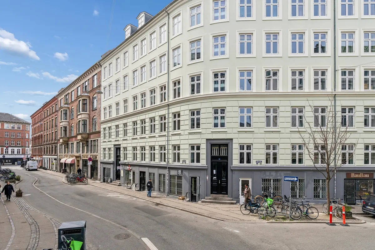 Forretningslokal med mange vinduer på Birkegade i København. Folk går rundt, og der er cykler parkeret foran. Solskin og blå himmel.