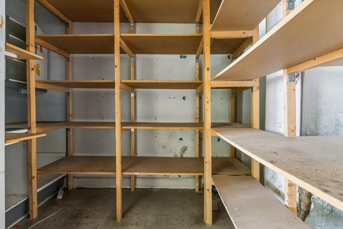 Store, tomme lagerhylder i nye lokaler med hvidt gulv og grå vægge med sletteforsøllinger og tærskler. Væggene er smuttede og mangler gulvbelægning.