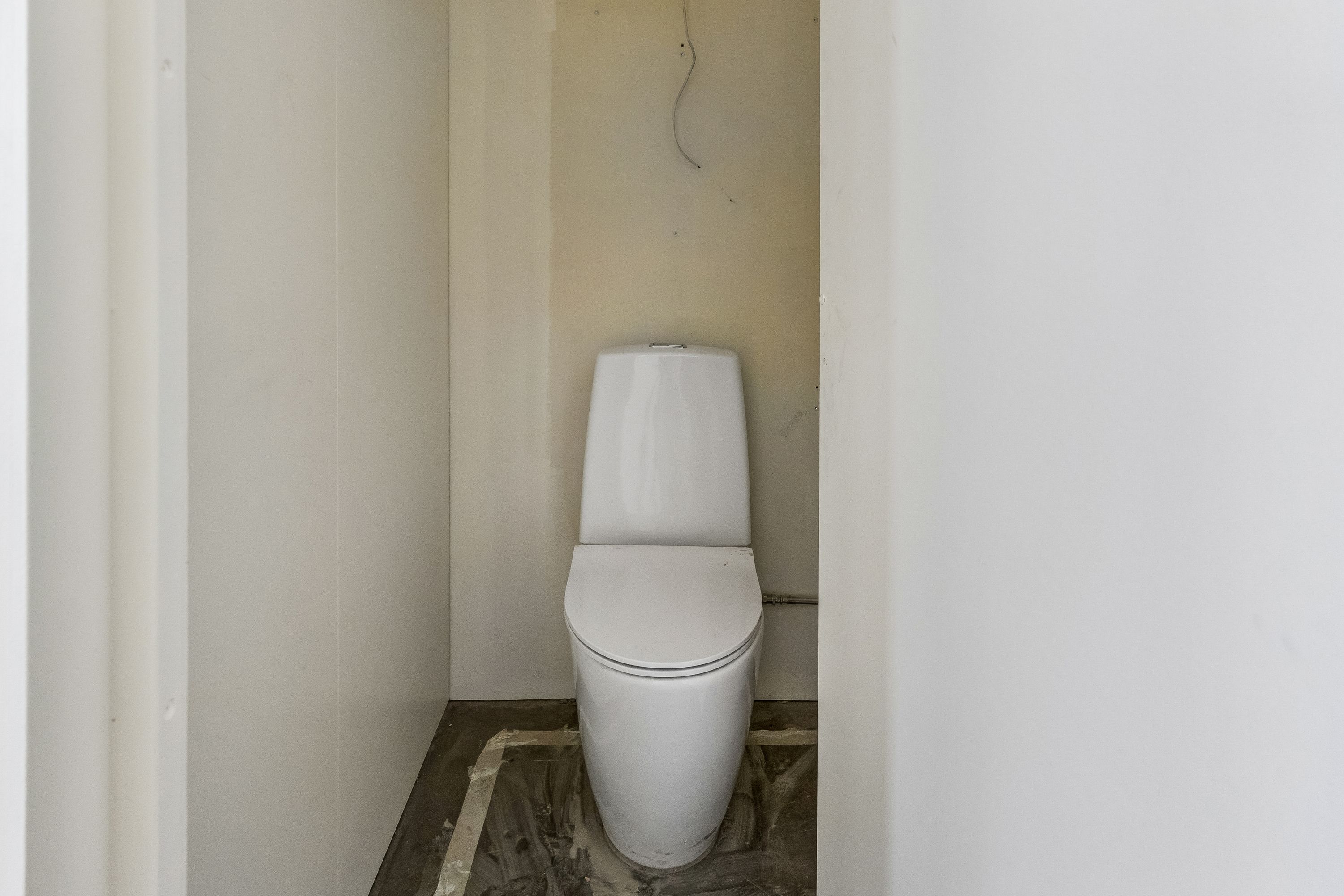 Et lille toiletrum med en hvid toilet i hjørnet mellem to vægge, der er malet hvidt. Toiletrummet er ubeboet og tømt