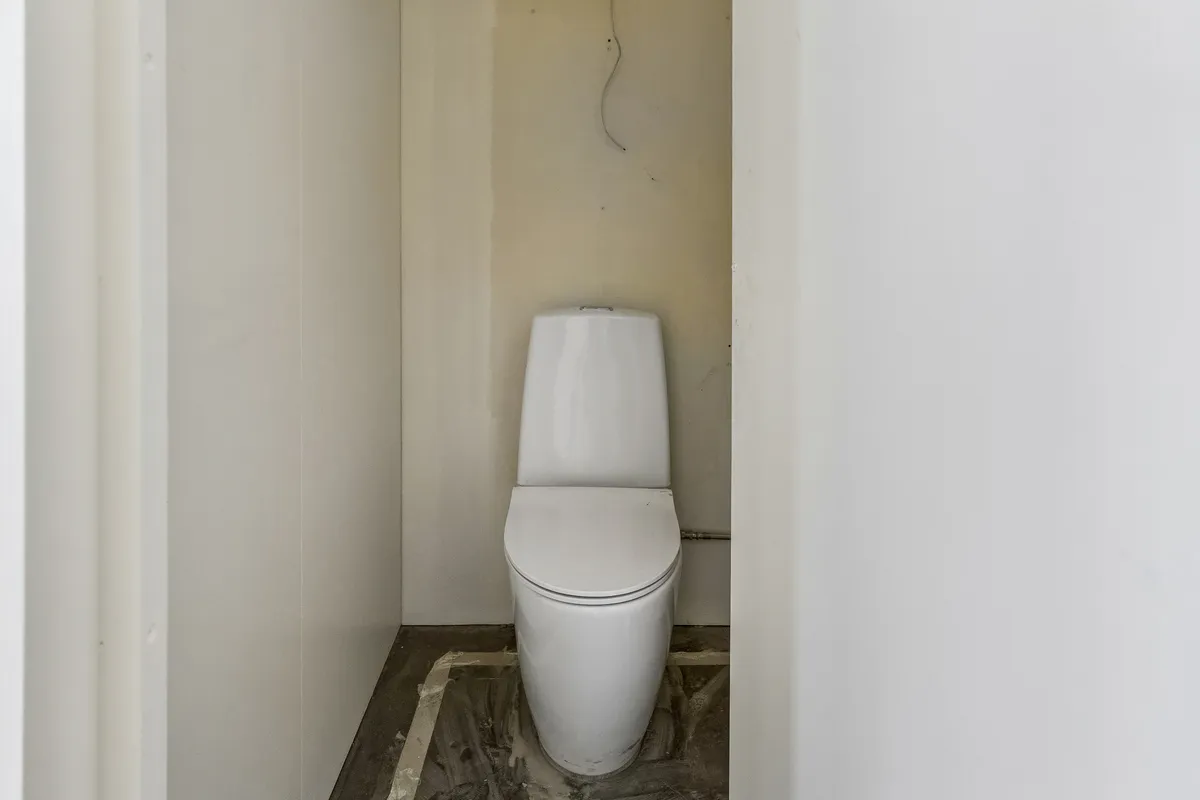 Et lille toiletrum med en hvid toilet i hjørnet mellem to vægge, der er malet hvidt. Toiletrummet er ubeboet og tømt