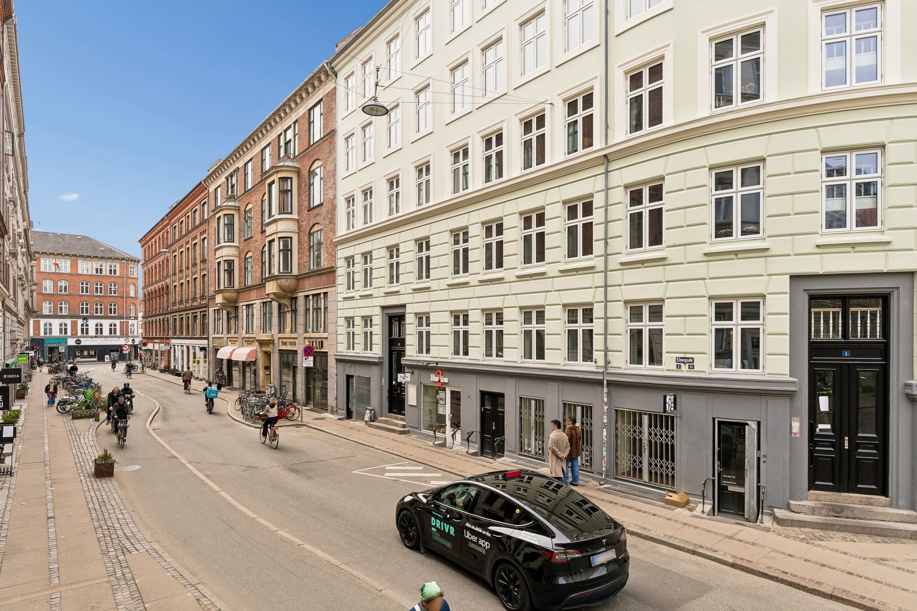 En bil kører på en gade med mange cyklister og fodgængere. Gaden er omgivet af store bygninger, og der er flere søjler og vægge med annoncer.