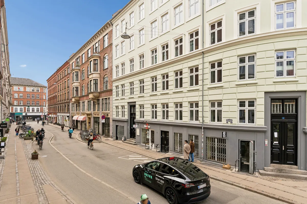 En bil kører på en gade med mange cyklister og fodgængere. Gaden er omgivet af store bygninger, og der er flere søjler og vægge med annoncer.