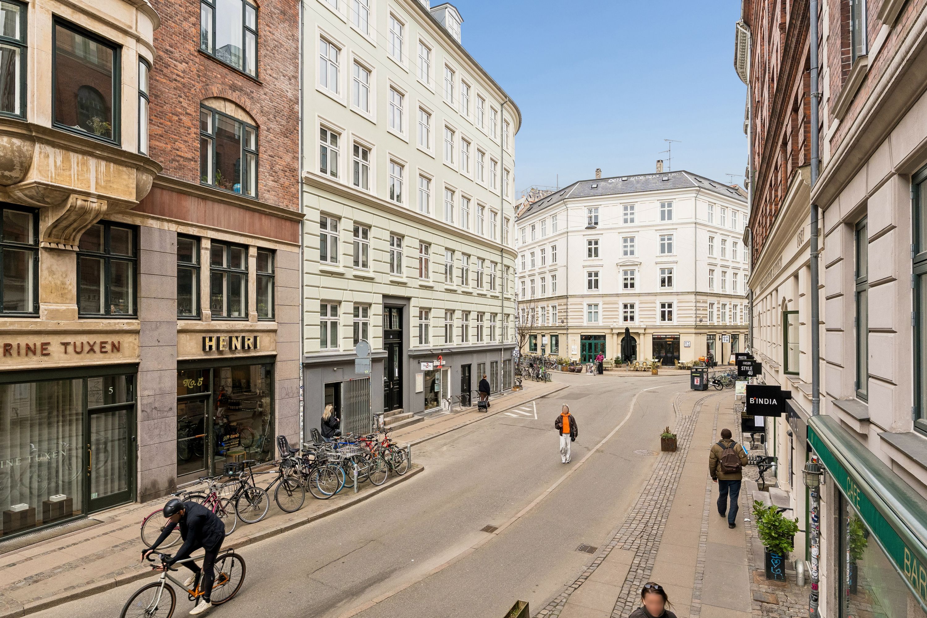 Cyklist kører på tom gade med butikker, hvor nogle er lukket, mens folk går på fortovet og tage er delvist dækket af skyer
