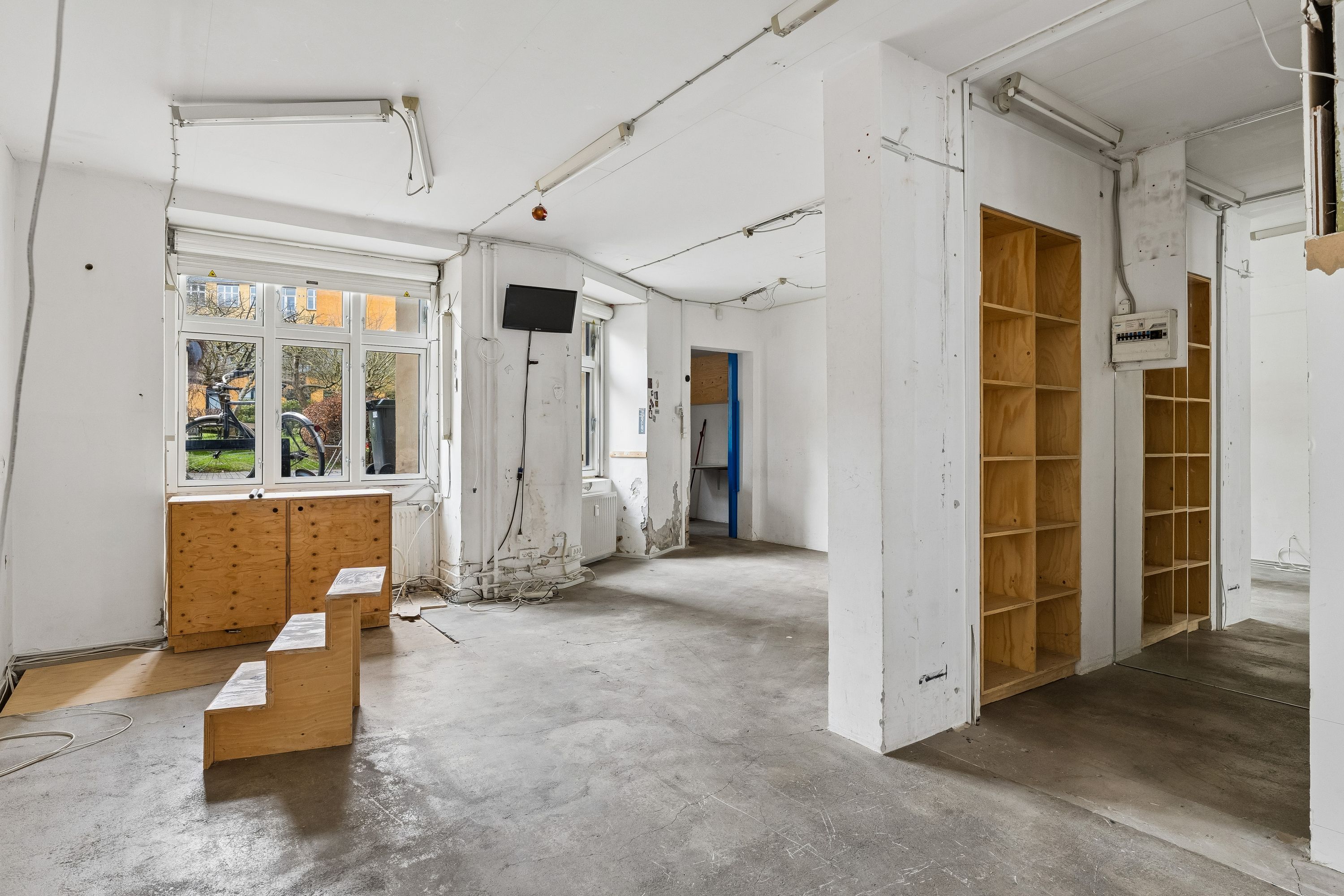 Tom rum med store vinduer, lysekroner og træhylder samt en tv-skærm og skraldespande udenfor