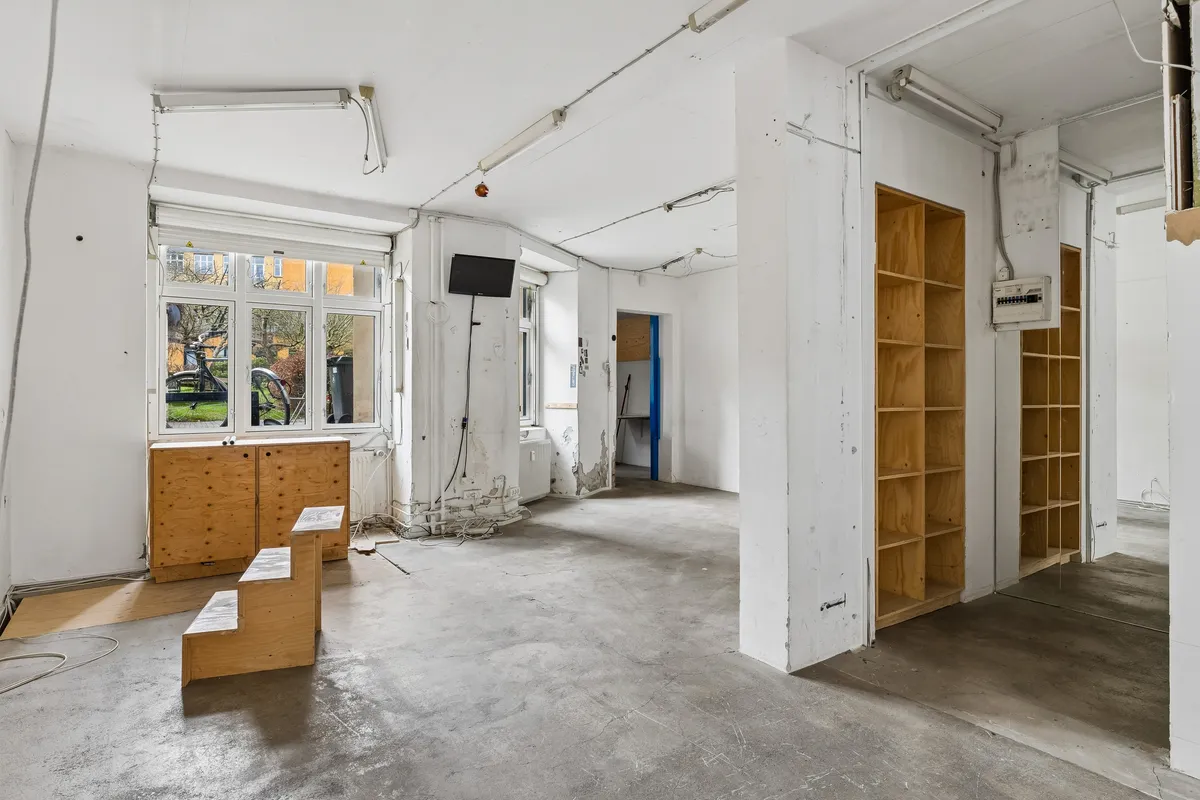 Tom rum med store vinduer, lysekroner og træhylder samt en tv-skærm og skraldespande udenfor