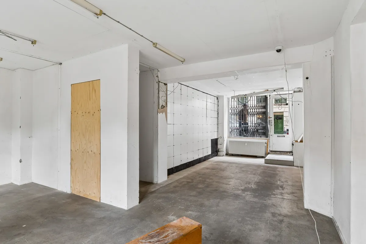 et tomt lokale med hvide vægge og lyse gulv. Der er et træbord og en trædør på den ene side. Der er også en dør og en radiator på den anden side