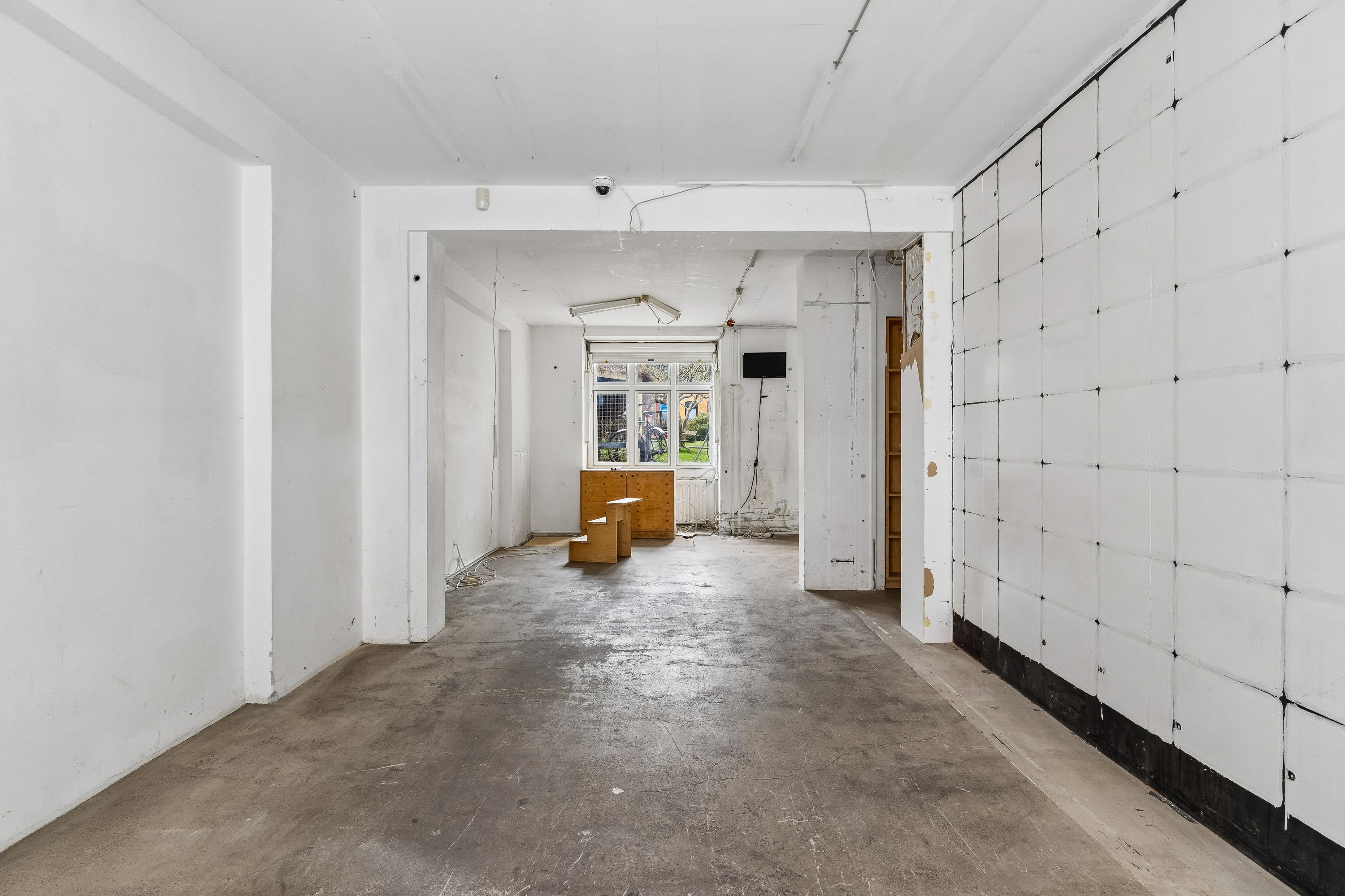 En ubenyttet og tømt lejlighedsrum med hvide vægge og cementgulv med to små trætrin og et skrivebord ved vinduet