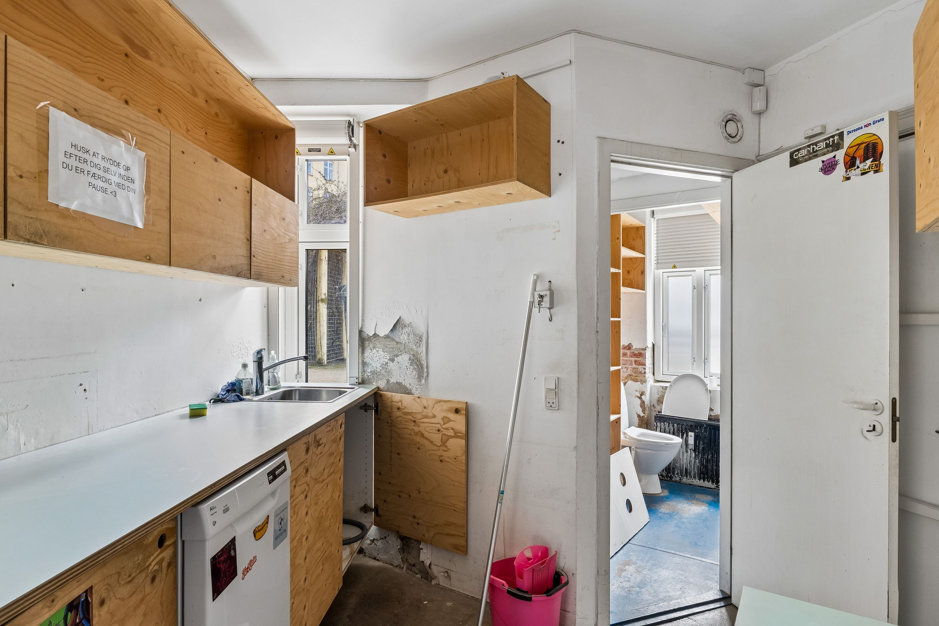 Lejlighed med køkken, vaskemaskine, åbent badeværelse og træhylder. Toilet med radiator og lys på loftet. Væg med hængende plakater og udkastet.