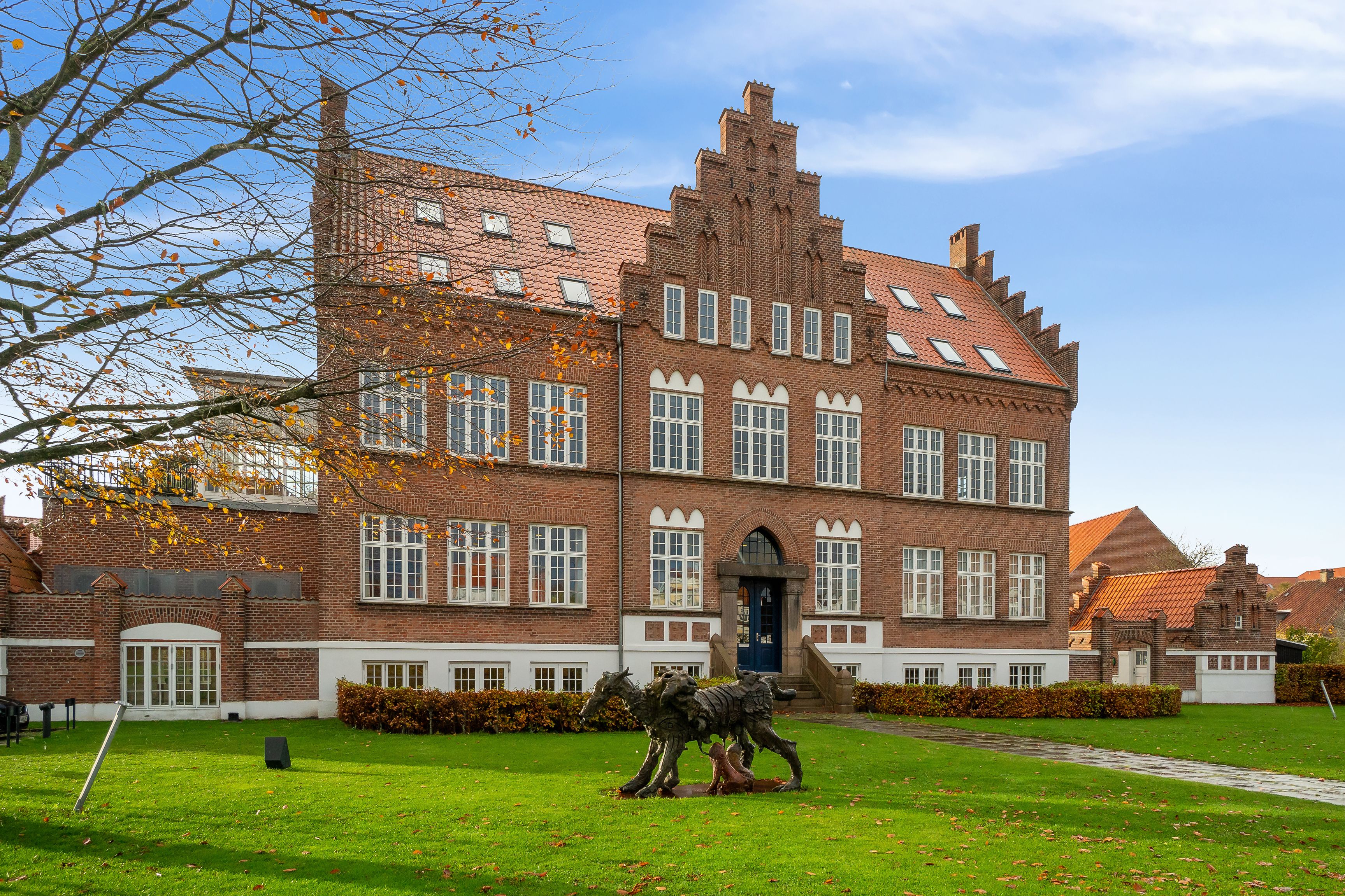 Store røde murstensbygning med terrasse og statuer foran, gul blomsterbed og brede vinduer på 3. sal
