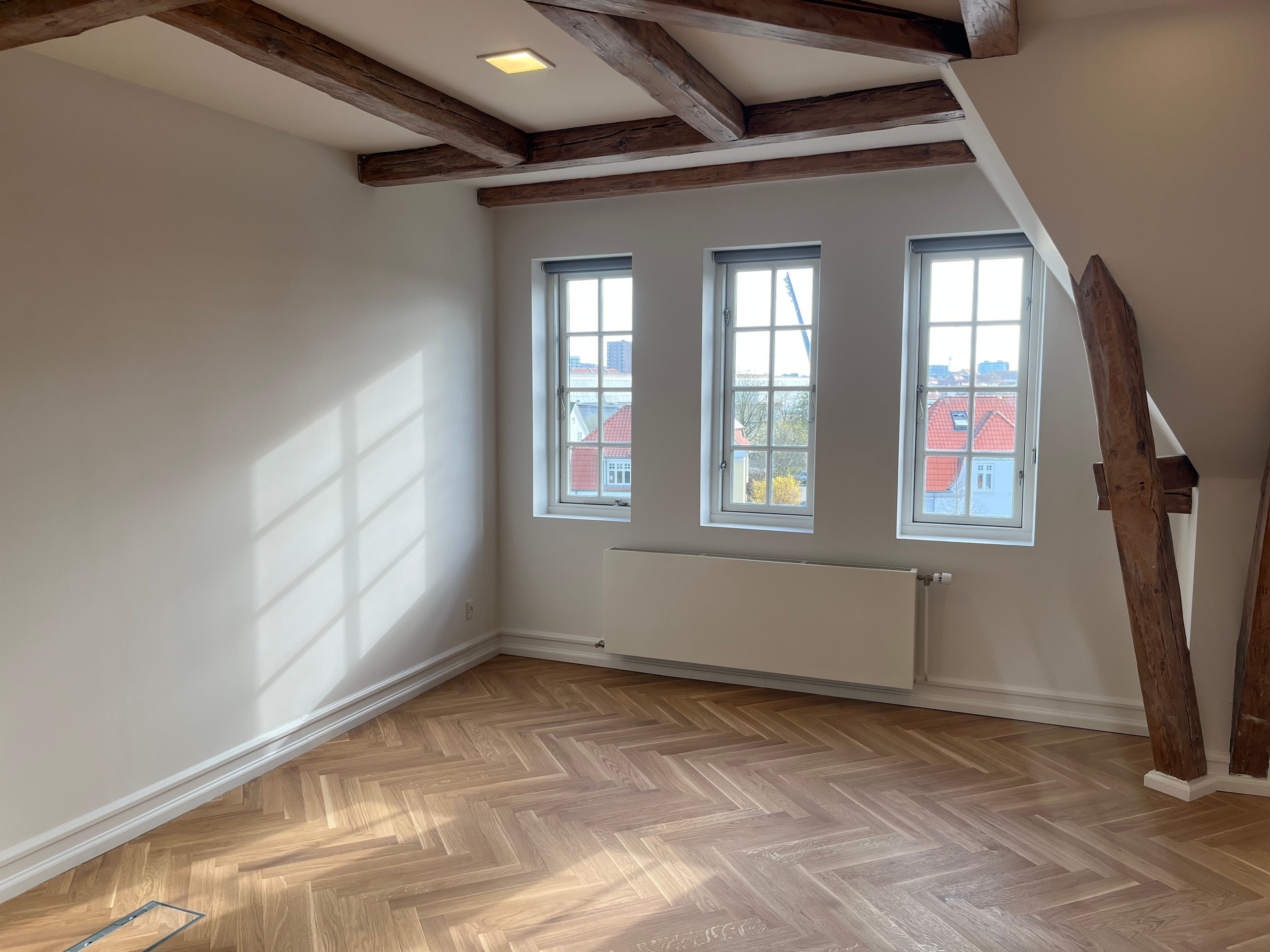 Solskin trænger ind gennem store vinduer i et stilfuldt loftsværelse med træbjælker og lysegrå vægge. En hvid radiator og trægulve gør det til et komfortabelt kontor eller arbejdsrum