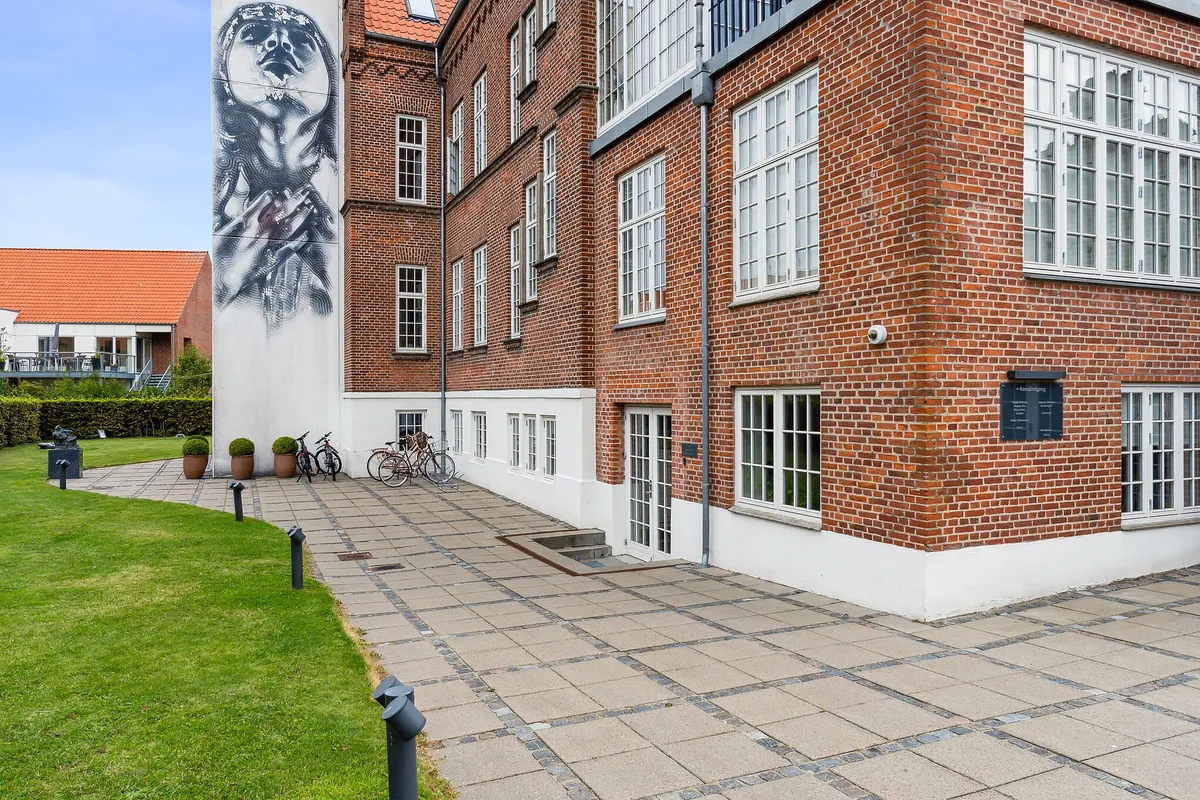 Store røde murstensbygning med vægmaleri og vinduer, to cykler og flere potteplanter, grøn plæne og gulvet er lavet af sten