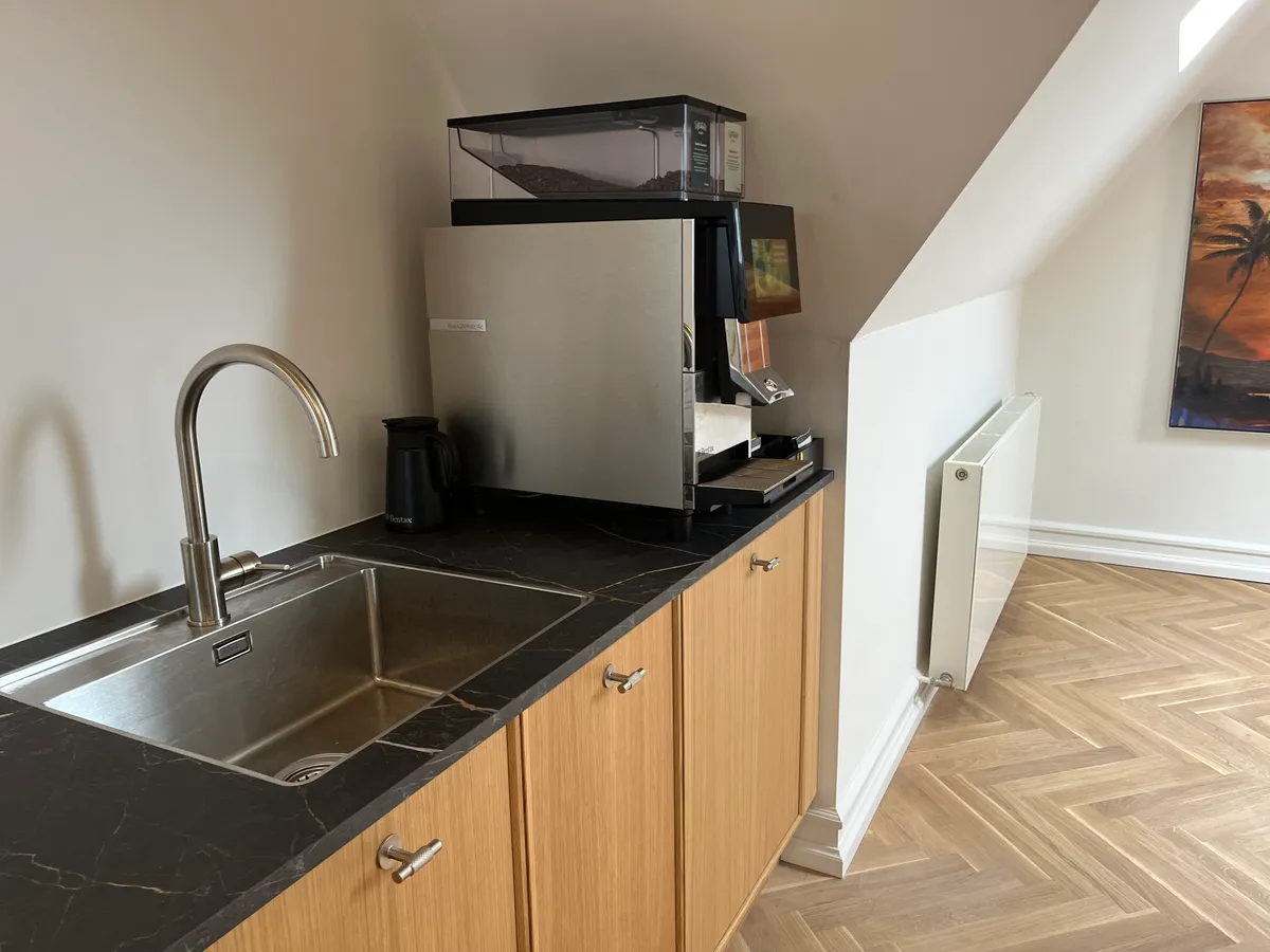 Billede af et moderne køkken med en stålvæg og trægulve. Væggen er malet hvidt og rummer en kunstmaleri. En stålvandhane med en sort kaffemaskine og en kaffemaskine med en kaffekværn ovenpå.