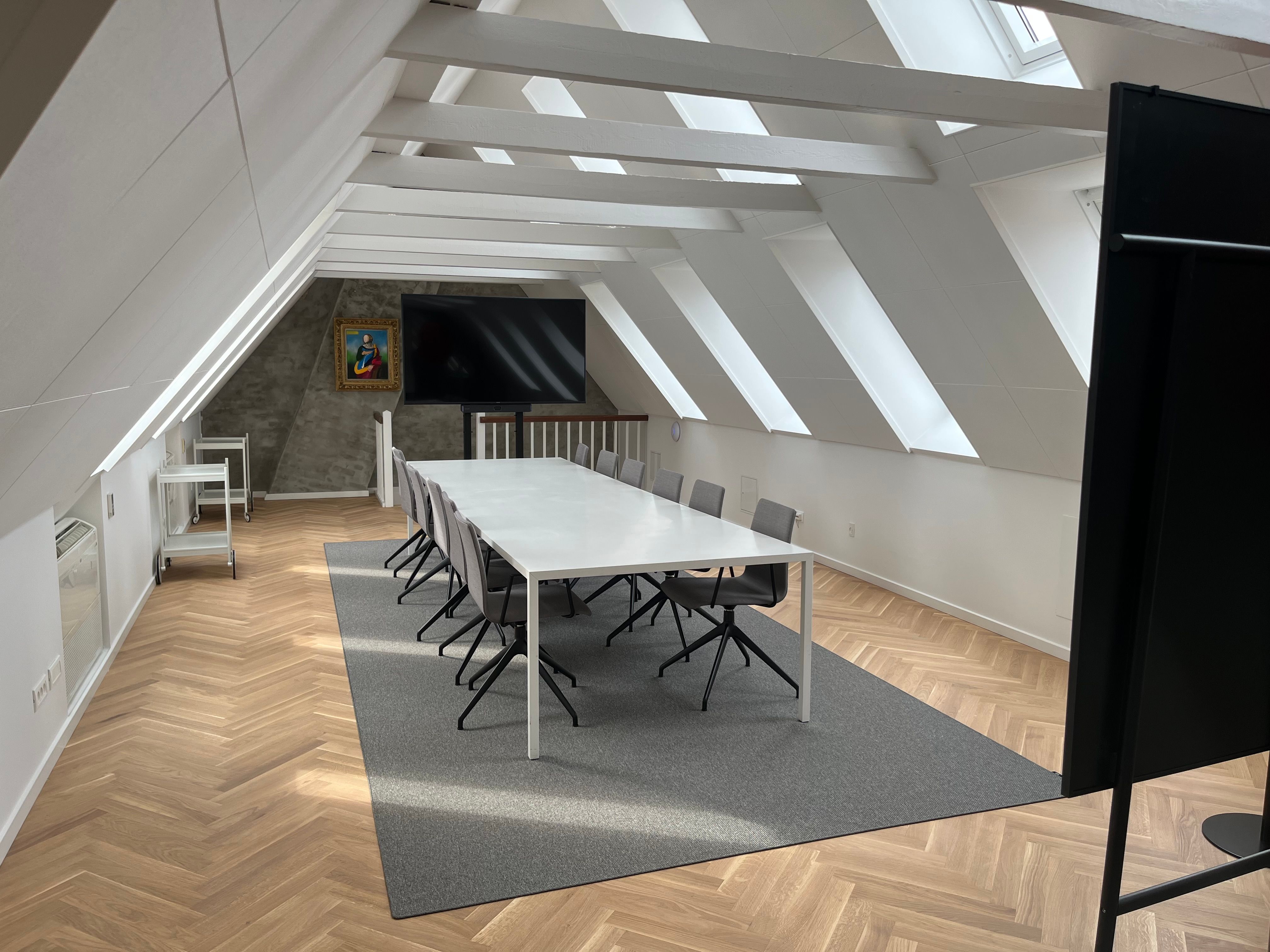 Loftsrum med hvid loftsbjælke, gulvtæppe og hvide møbler, herunder en lang konferencebord med stole, og en tv-skærm