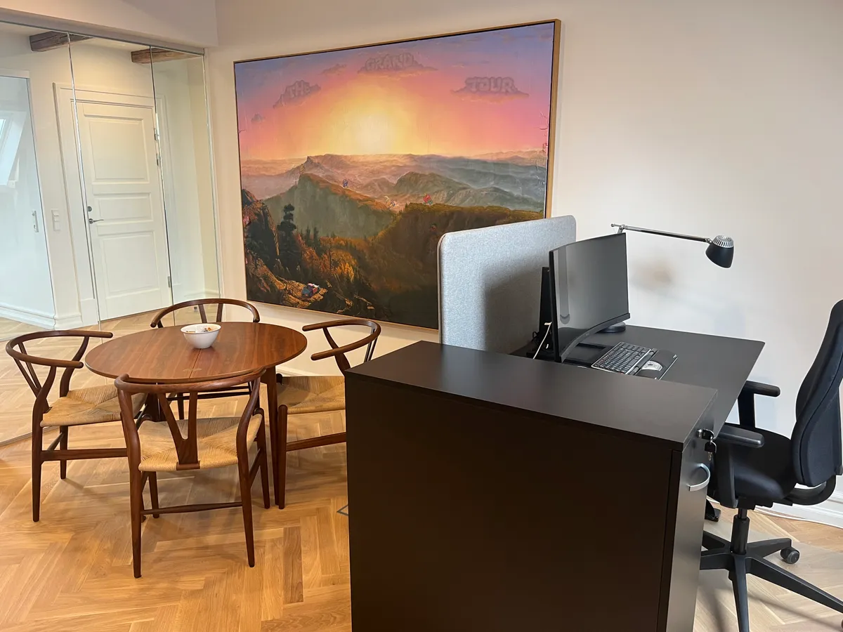 Hjørnekontor med skrivebord, stol og runde bord med skål. Maleri med bjerge og solnedgang på væggen. Glasdør og højloft i baggrunden.