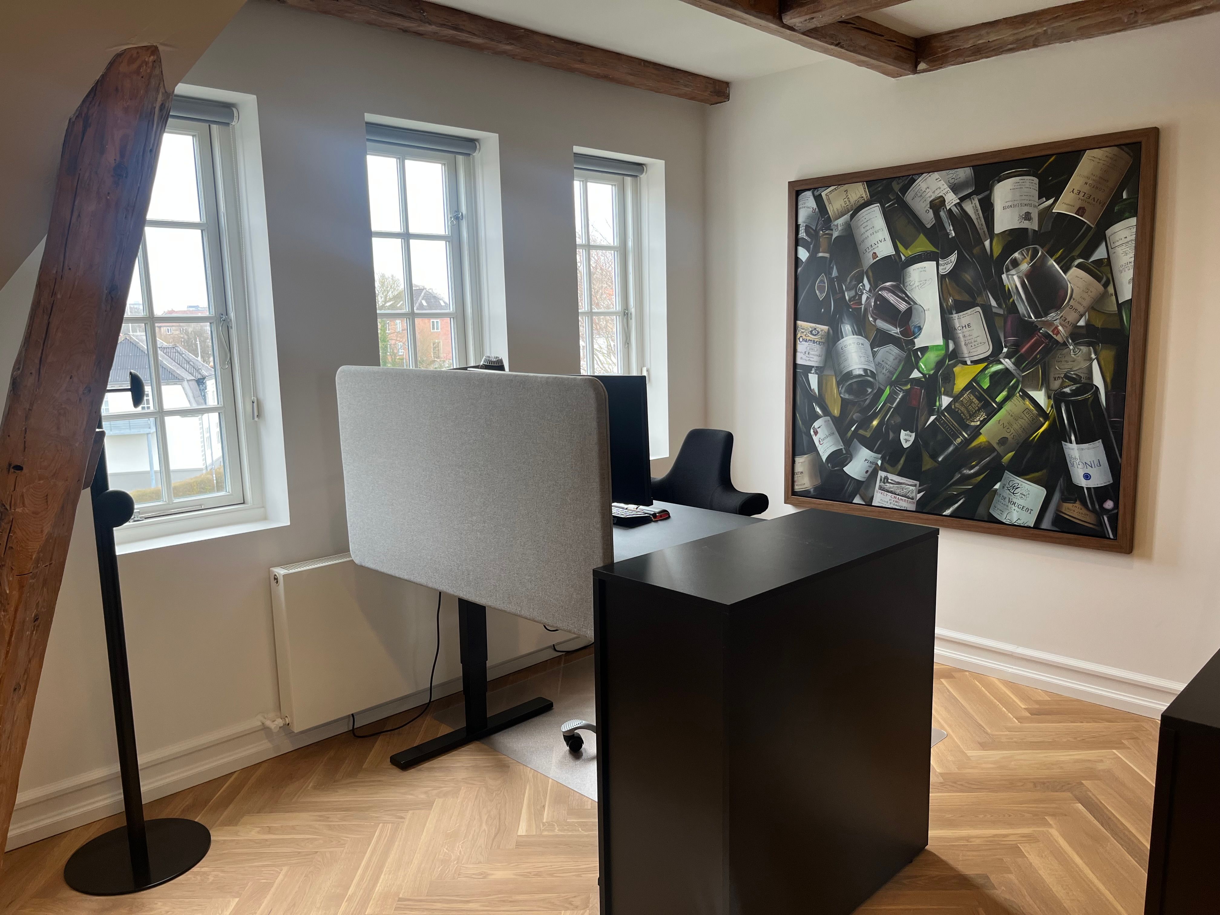 Kontor i Aalborg med skrivebord, stol, højttalere, gulv og maleri på væggen. Væggen er hvid og loftet er lavet af træ.