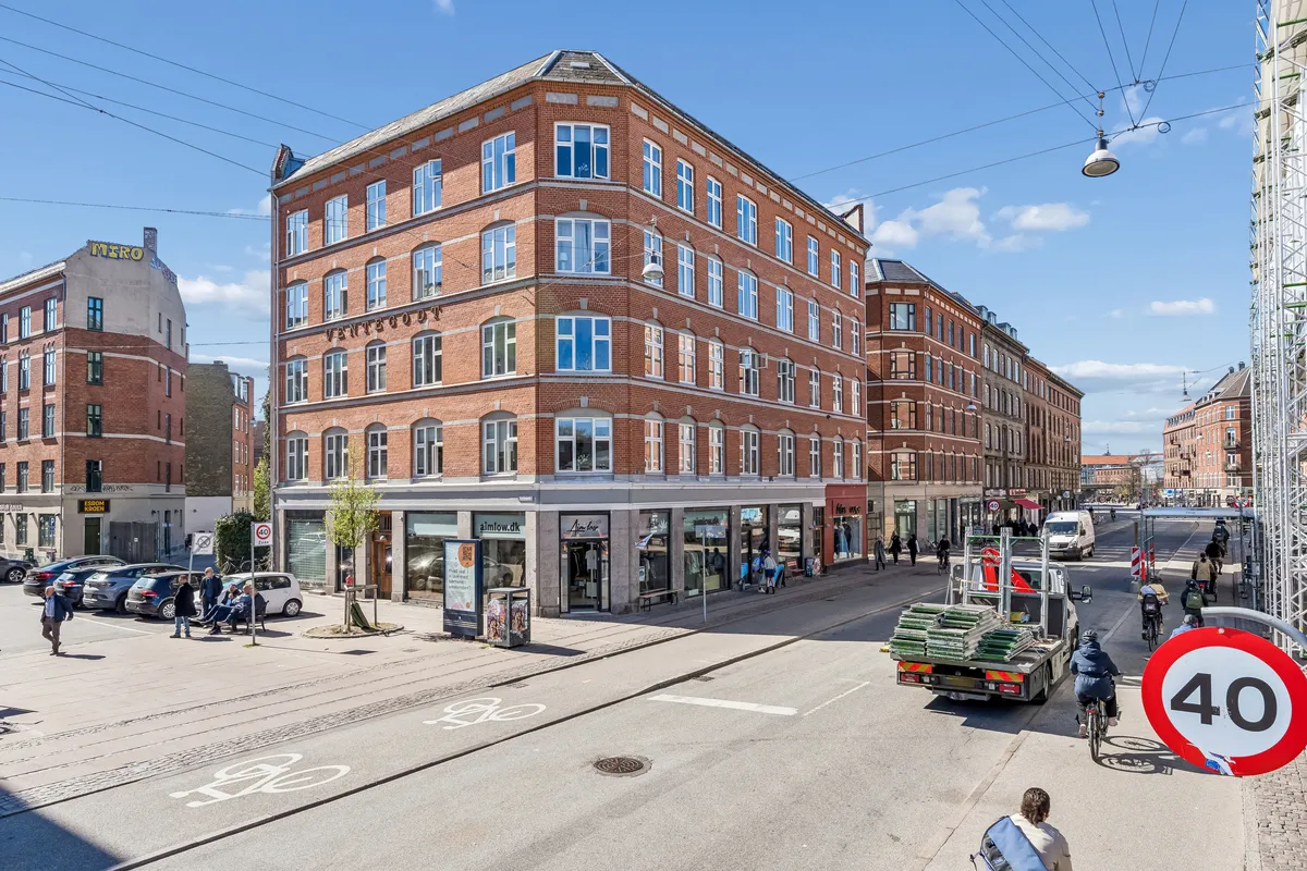 Butik til leje på Nørrebrogade i København N. Mennesker cykler og går på gågaderne og parkeringspladser. En cykel og en bil passerer gennem et 40 km/t-område.