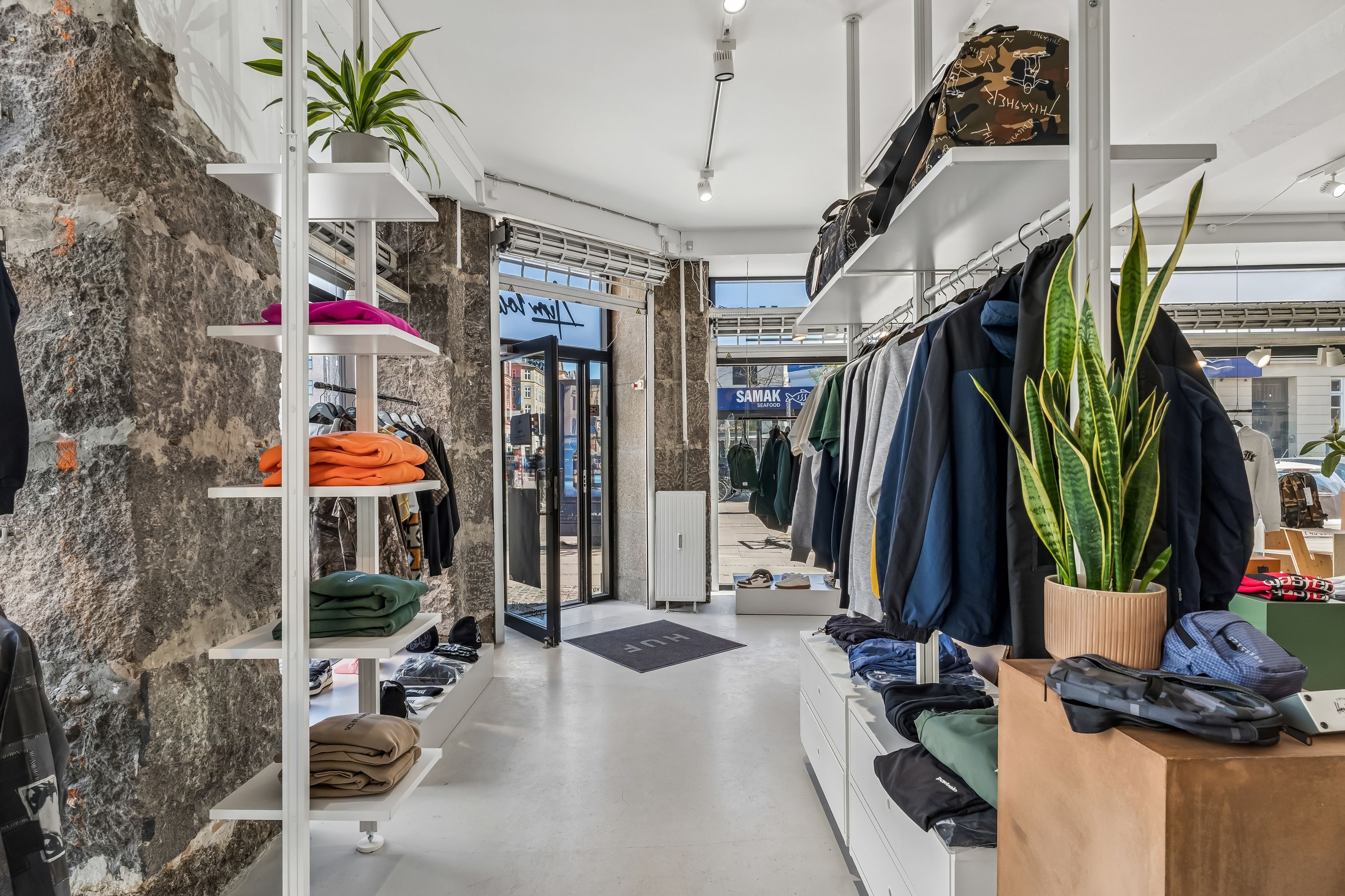 Butik med klædtøj og sko i hvidt og blåt og to store gulvvægge med grønne planter i lysebrune potter og lysekroner på loftet