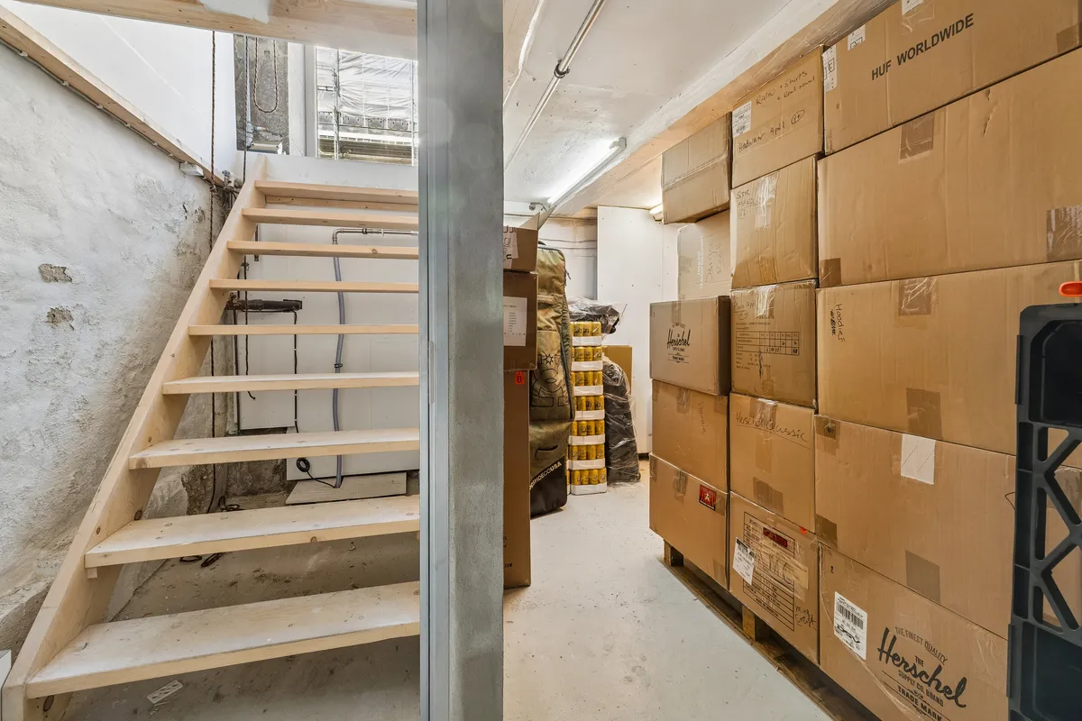 Foto af et lager med forskellige brunæ kartoner og et trappeforløb. Væggene er gule og rå, og der er en køleskab med ølflasker