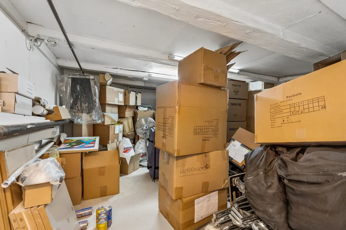 Lagerrum med mange kartoner, plastasker, emballage og en ventilator i hjørnet, samt en række skriftligt mærkede bokse med varer og en klippe.