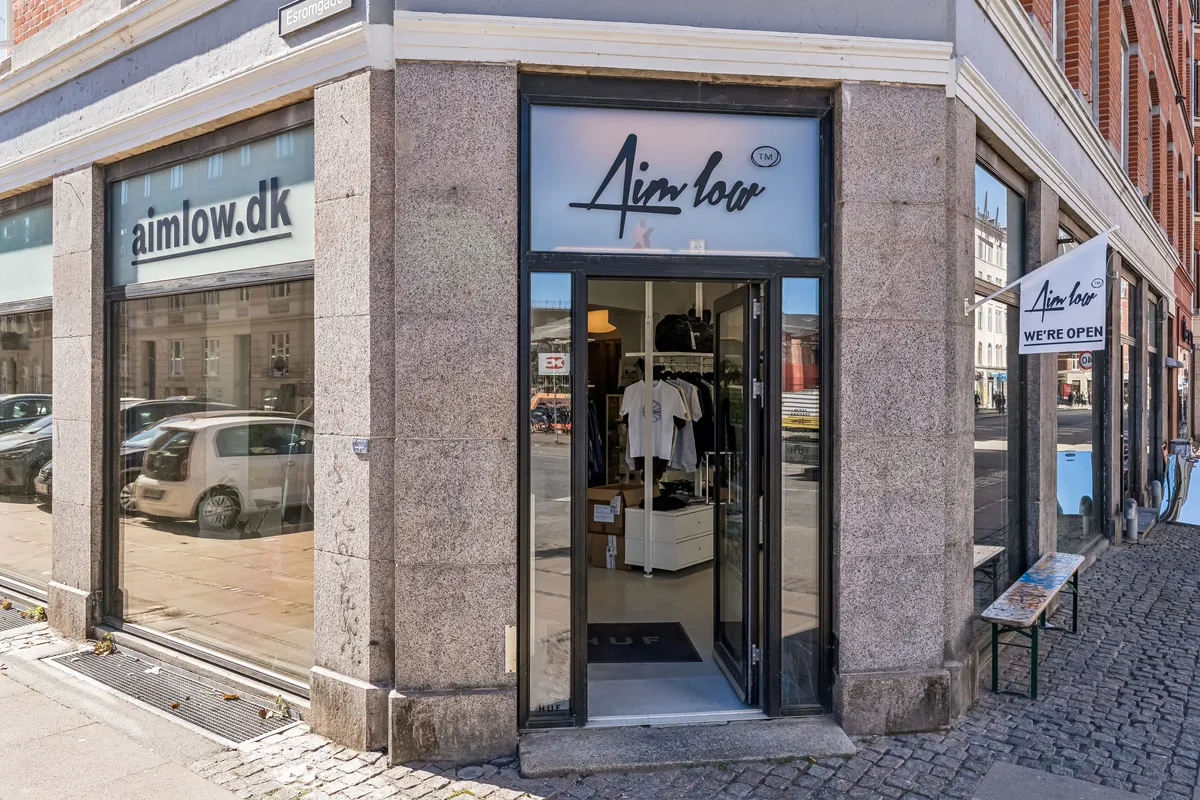 Butikslokale med glasvinduer og en åben dør, skrift Aim Low og WE'RE OPEN hænger udenfor, tøj og sko står i udstykningen, hvid banner med tekst Aim Low og WE'RE OPEN