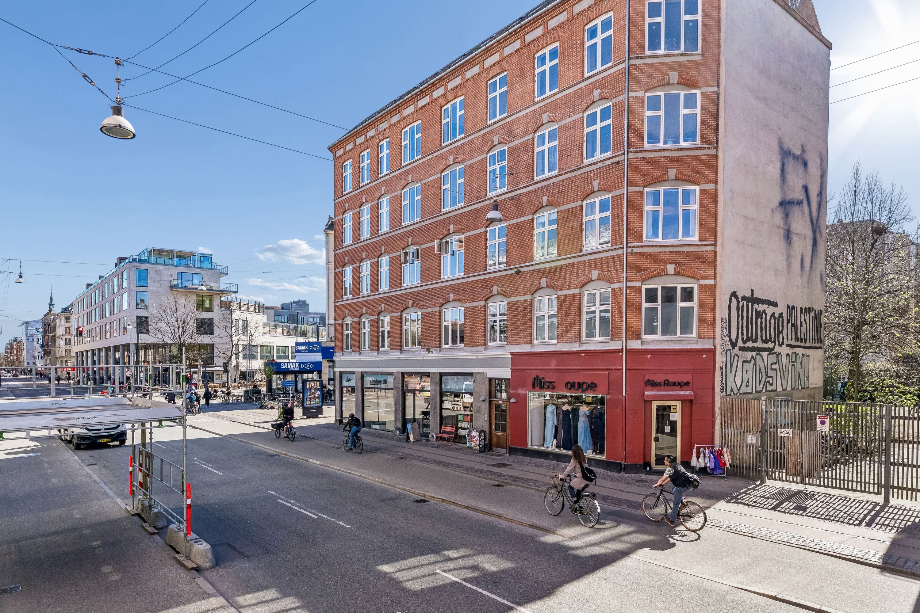 Mennesker cykler og går på gågaden med butikker og graffiti på bygninger. Bygning med røde facader og butik Miss Ouge. Personer bærer tasker og reder cykler.