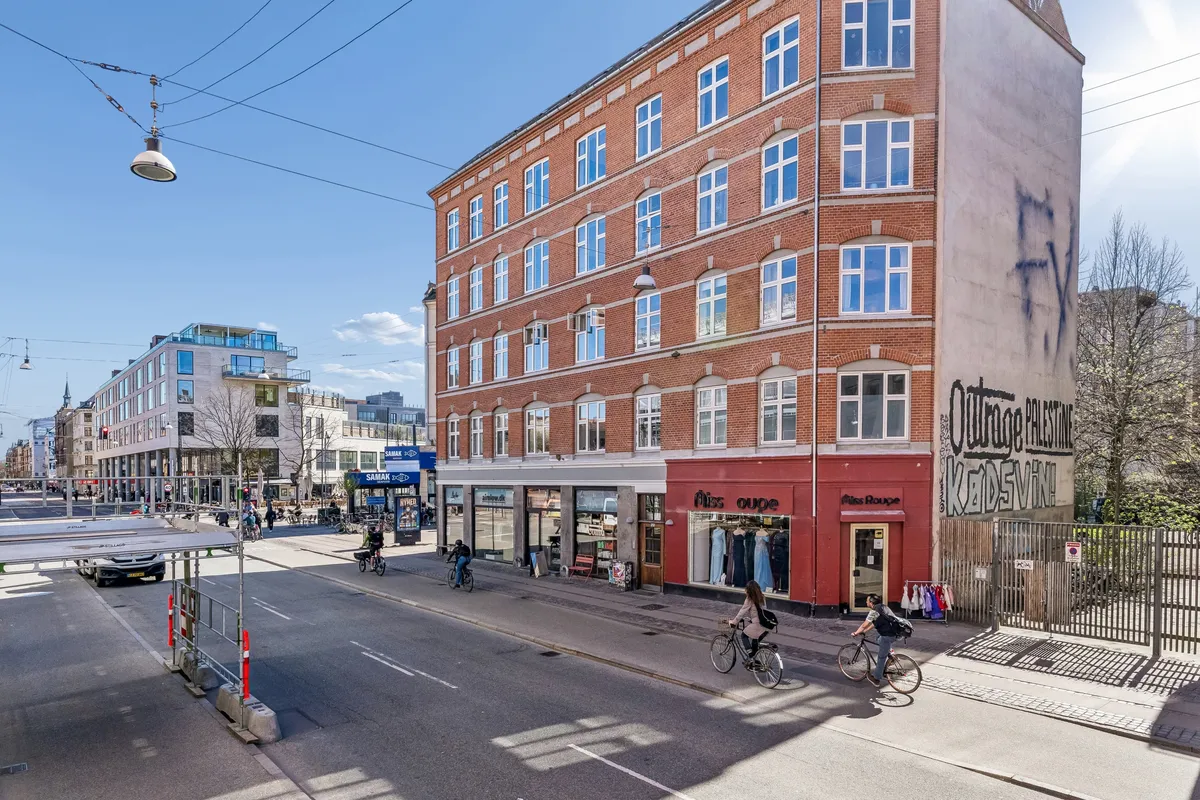 Mennesker cykler og går på gågaden med butikker og graffiti på bygninger. Bygning med røde facader og butik Miss Ouge. Personer bærer tasker og reder cykler.